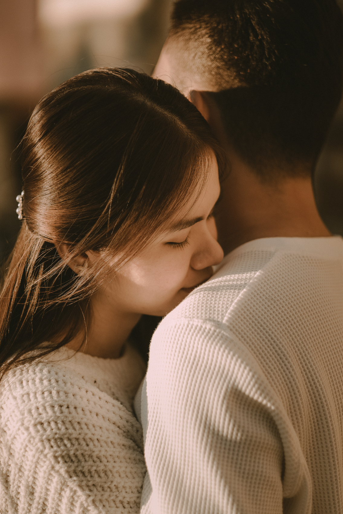 A young woman with long dark hair and a young man with short hair hugging closely, with her eyes closed and her face resting against his shoulder.