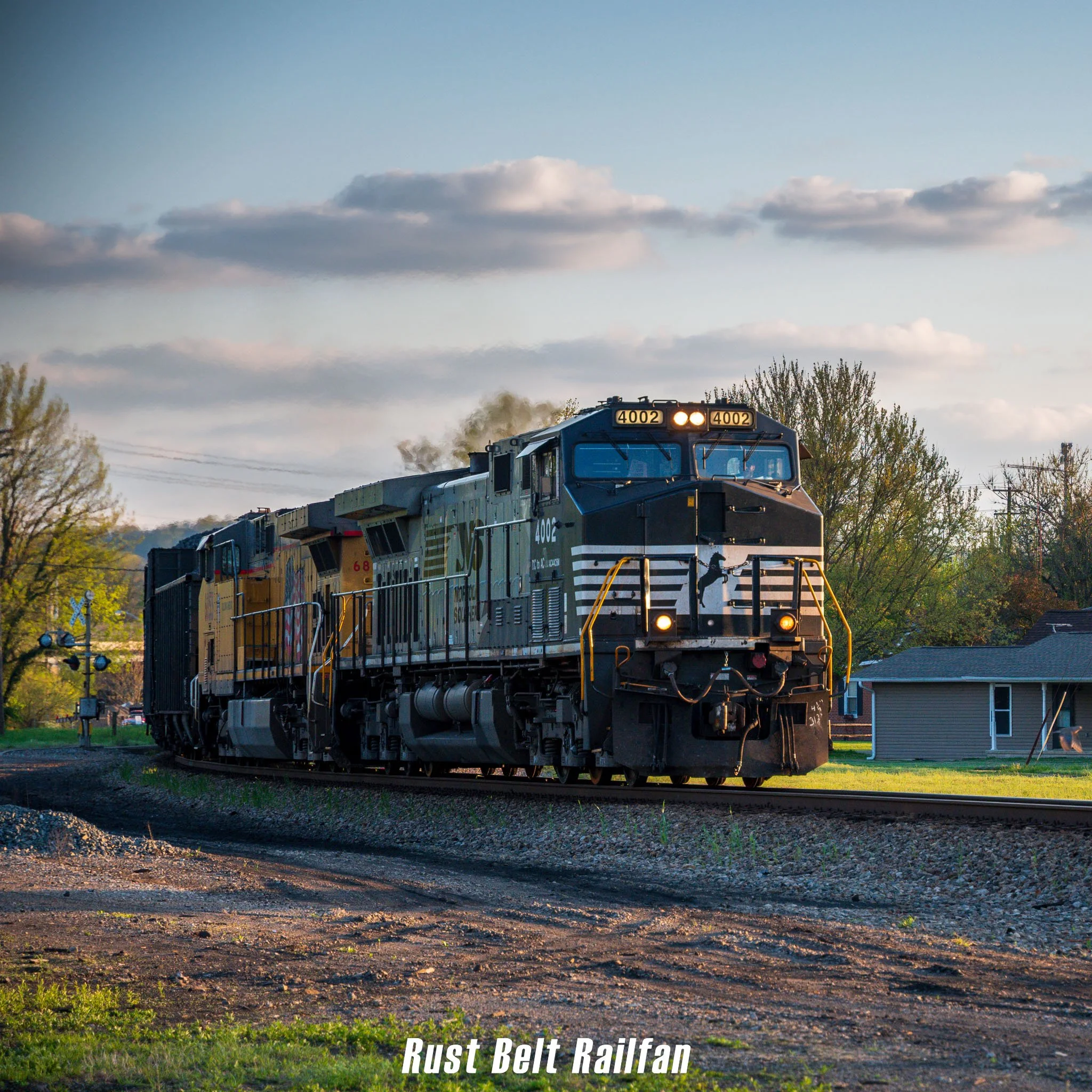 A black and gray diesel train moving on a railroad track in a suburban area during sunset, with trees, houses, and a clear sky with clouds in the background.