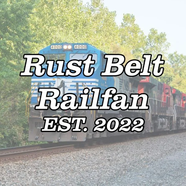 Blue and black locomotive train moving on railway tracks with trees in the background. Overlaid text reads: "Rust Belt Railfan EST. 2022."