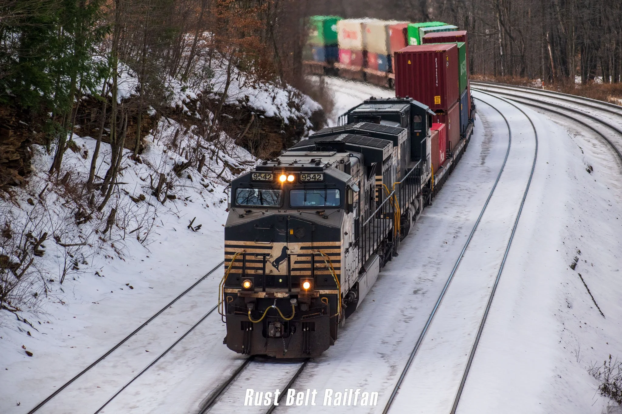 A freight train traveling through a snowy wooded landscape, viewed from an elevated angle.
