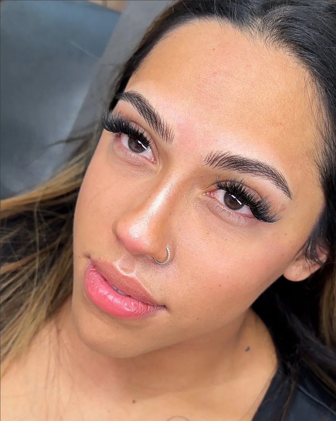 Close-up of a woman with long, thick eyelashes, brown eyes, and a nose ring. She has light makeup and a small mole on her neck.
