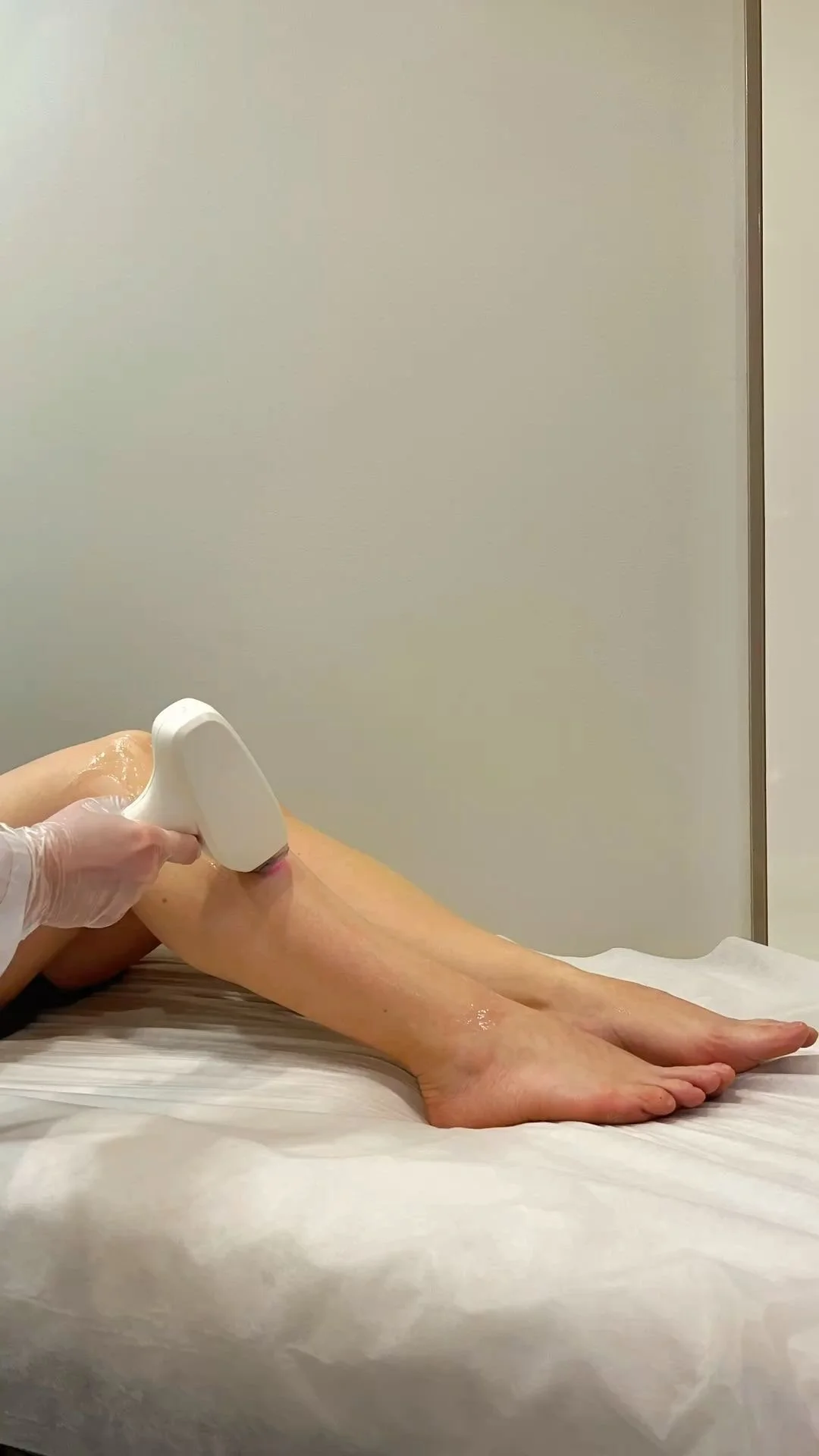 A person's leg being treated with a handheld device, possibly for a medical or cosmetic procedure, on a massage table in a clinical setting.