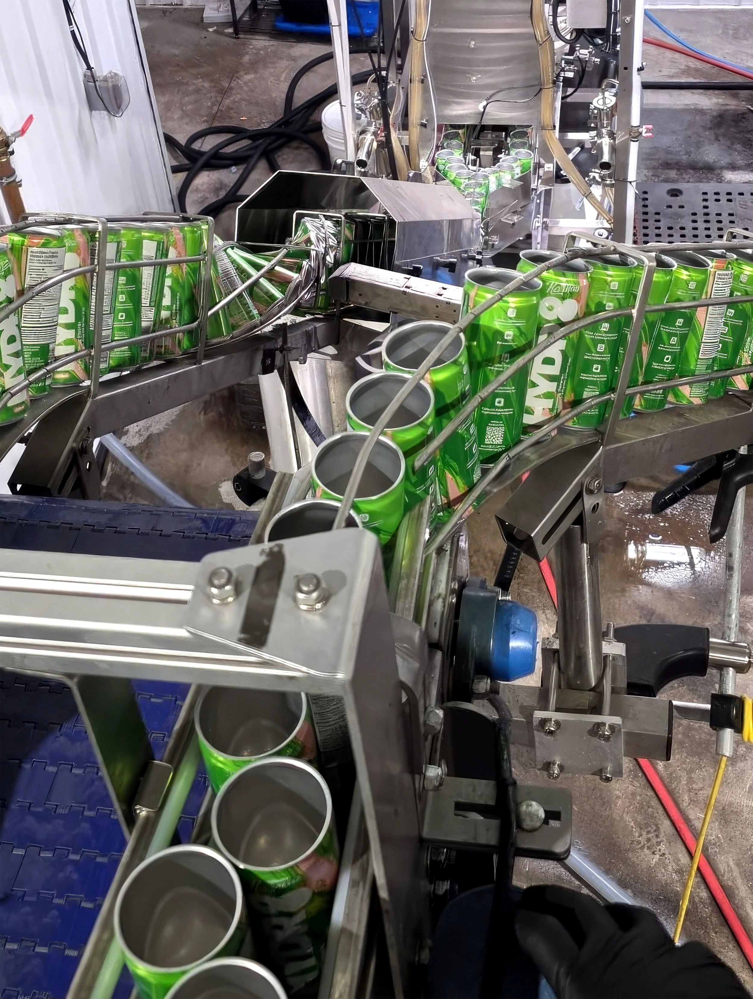 A manufacturing or bottling facility showing a conveyor belt with green aluminum cans being processed. The background has industrial equipment, pipes, and wires.