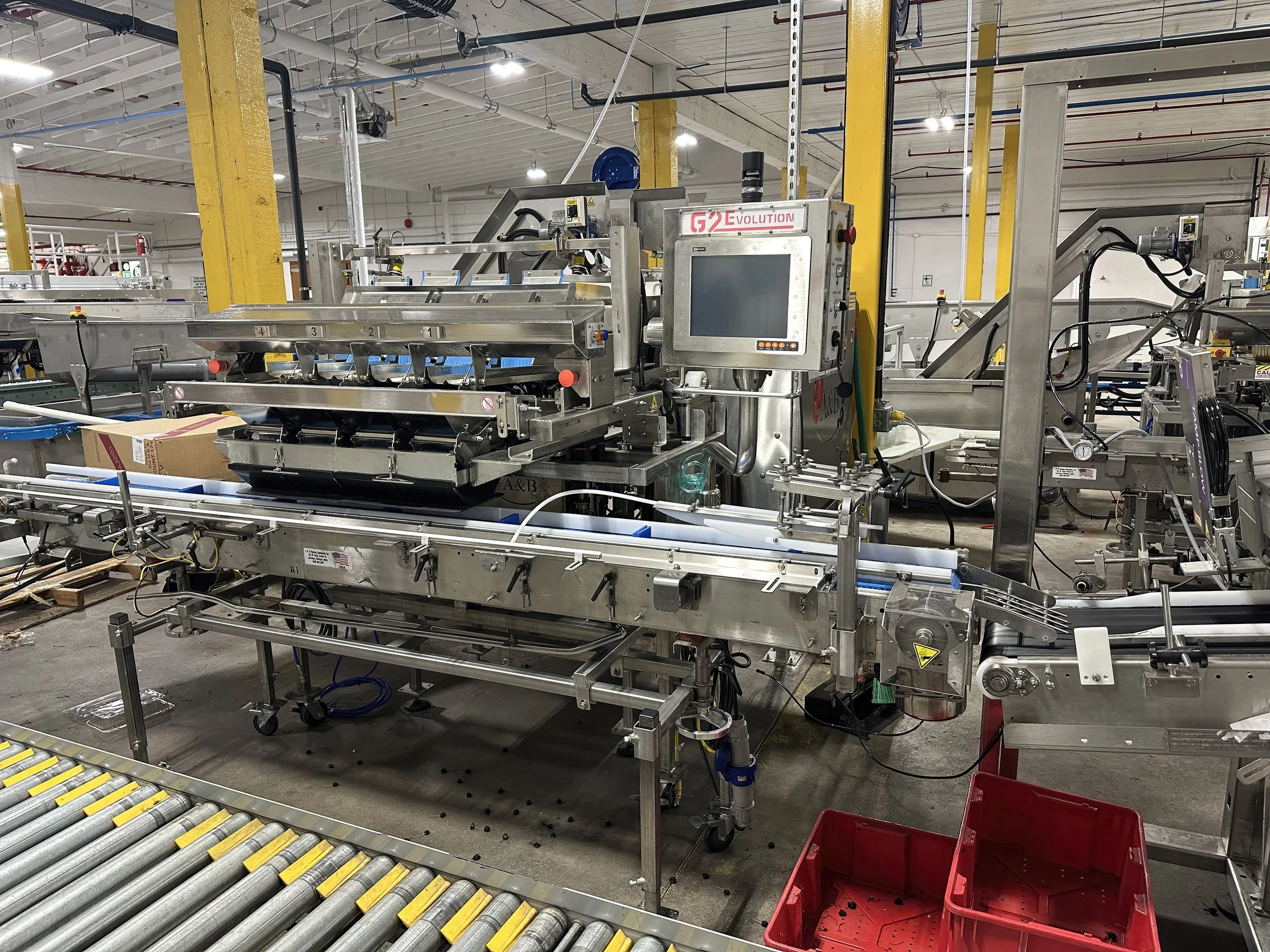 Industrial conveyor system with machinery in a warehouse, surrounded by yellow support pillars and various equipment.