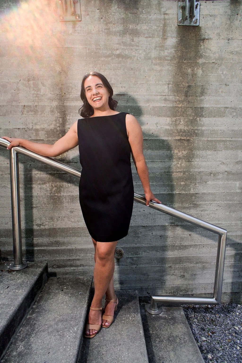 A woman in a black sleeveless dress and beige high heels standing on a concrete staircase with her right hand on the metal railing, smiling and looking slightly to her left, against a concrete wall background.
