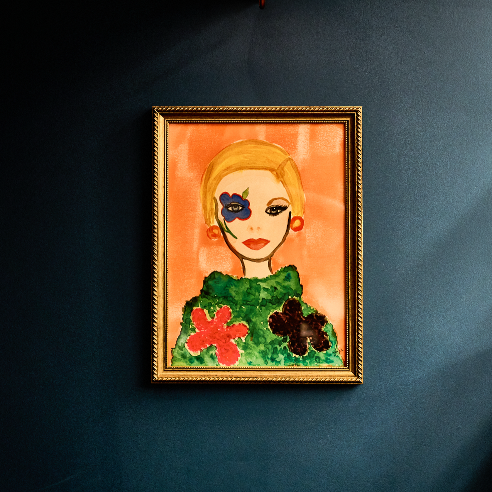 Retrato pintado de uma mulher com cabelo loiro, usando uma roupa verde com flor vermelha e preta, com máscara de pintura azul e detalhes em verde ao redor do olho esquerdo, em uma moldura dourada, pendurado em uma parede azul escura.