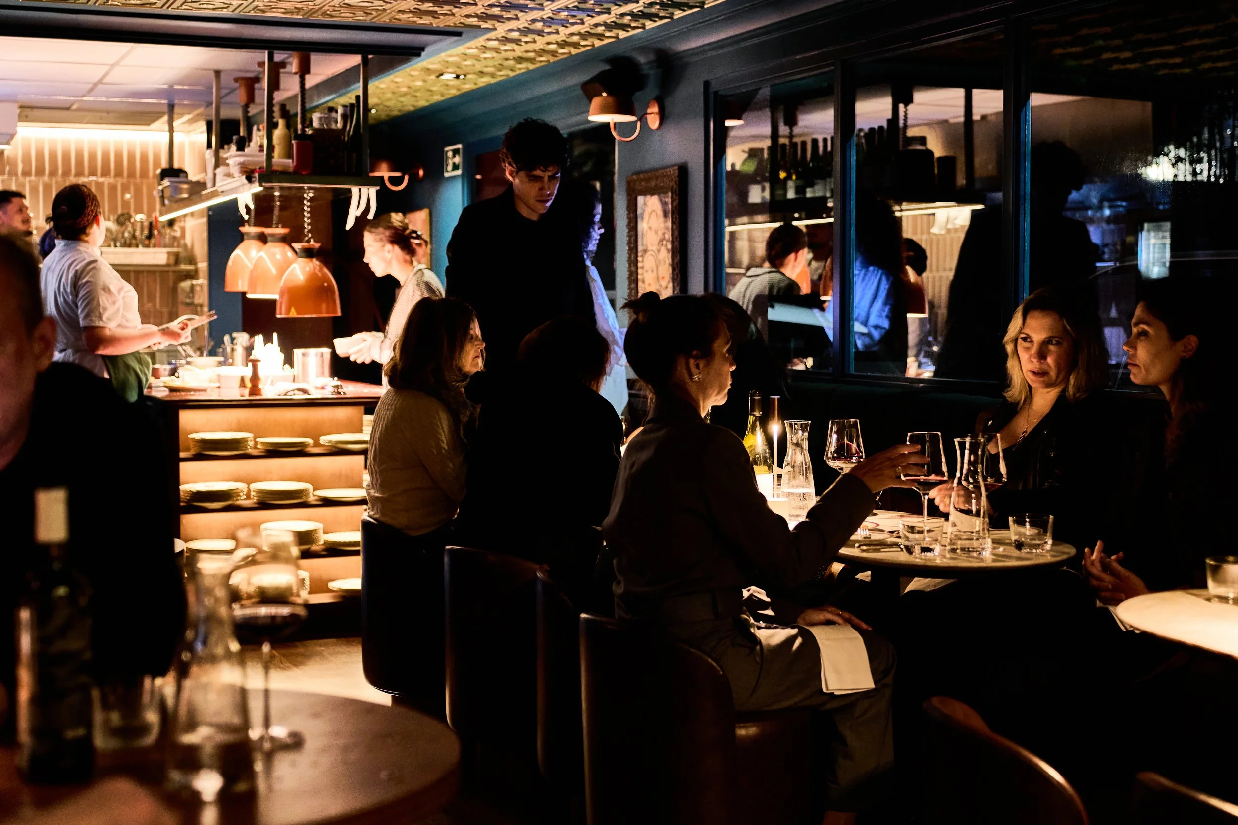 Pessoas jantando em um restaurante elegante à noite, com uma cozinha visível ao fundo.
