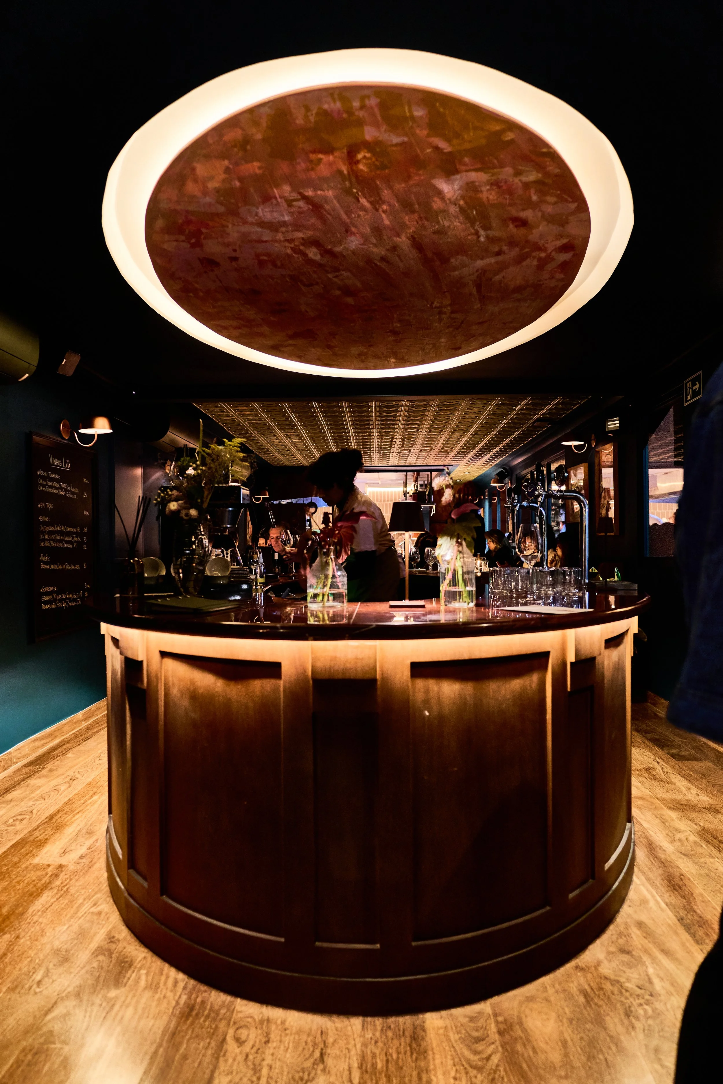 Bar com teto circular com luz, vasos com flores na bancada de madeira escura, pessoas ao fundo em um ambiente com iluminação fraca.