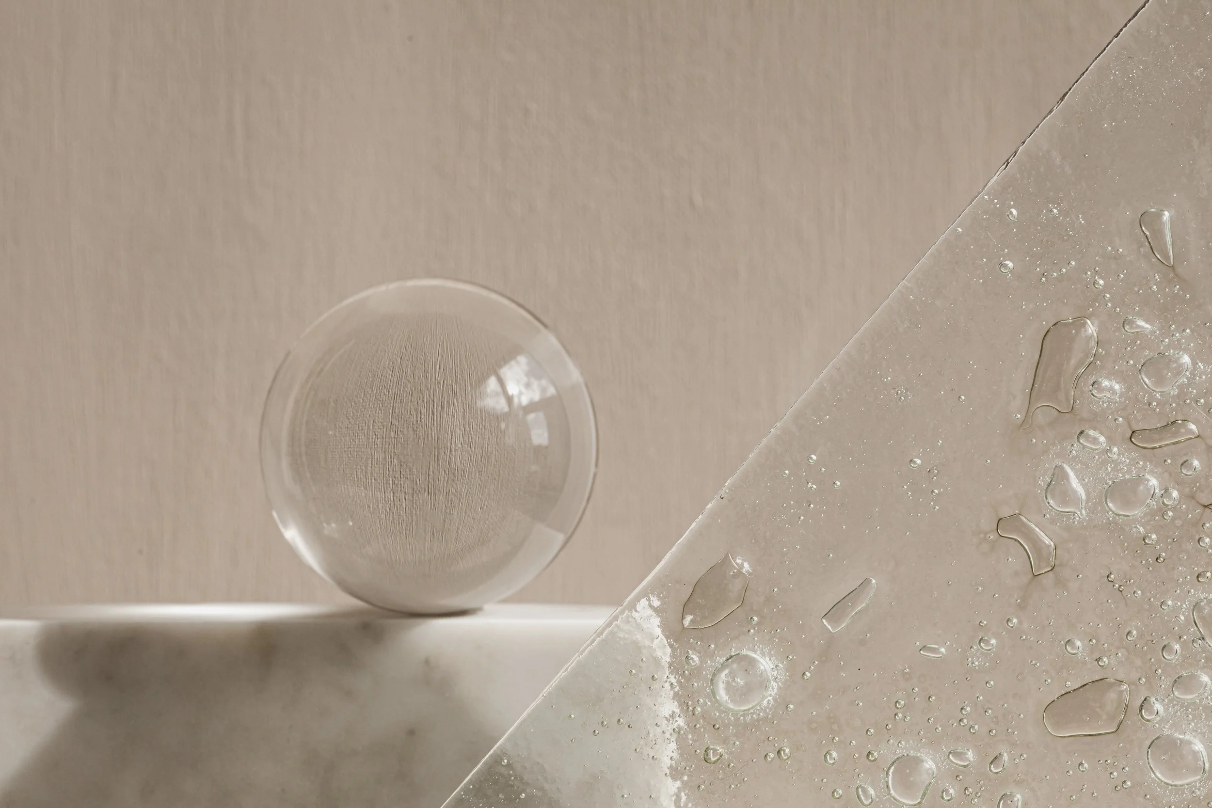 A glass sphere resting on a marble surface, with a textured beige wall in the background. The image is divided diagonally to show a close-up of water droplets on the marble surface.