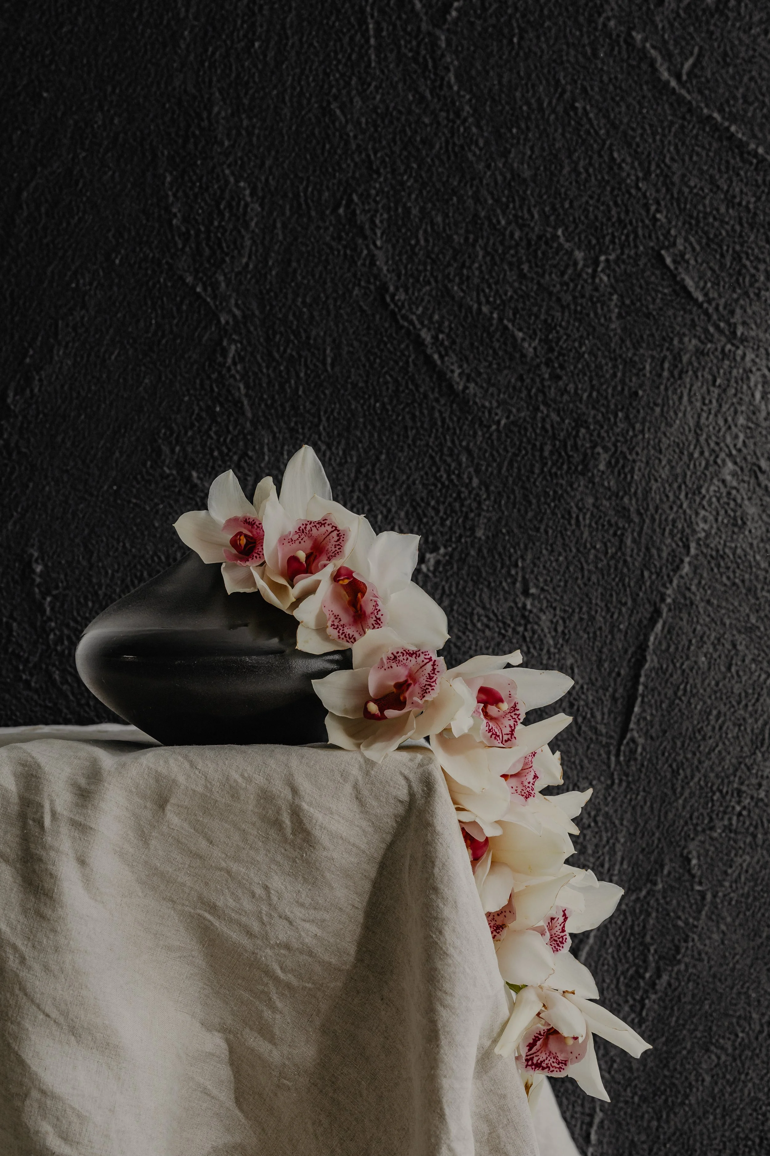 A black vase filled with white orchids with pink and red markings, placed on a beige fabric-covered surface against a dark textured wall.