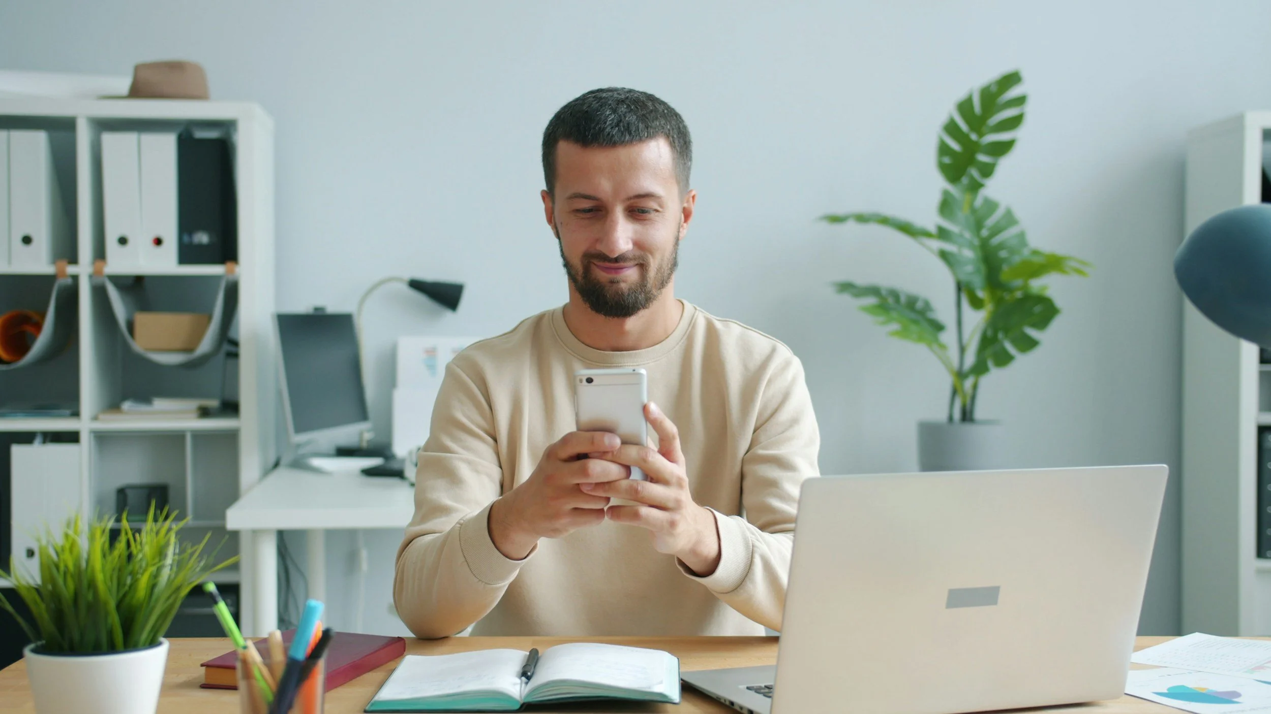A man with a beard and short dark hair sitting at a desk in an office, looking at his smartphone and smiling.