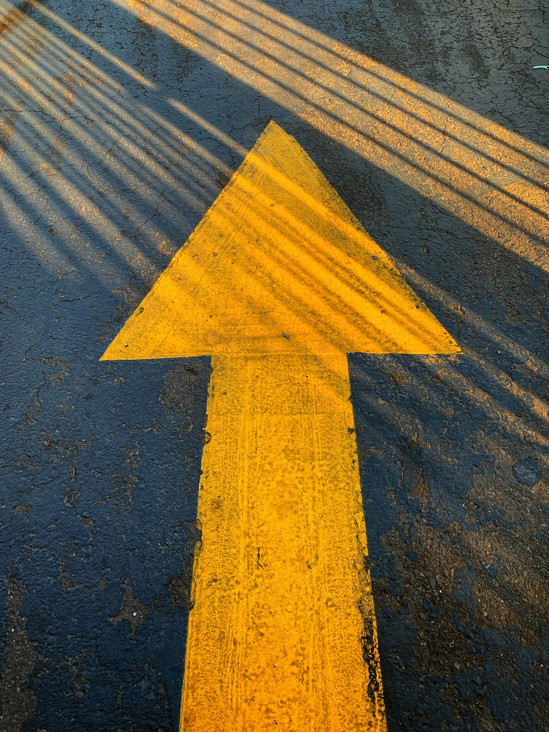 A yellow arrow painted on a dark, cracked asphalt road road with tire marks and shadows of a nearby fence.