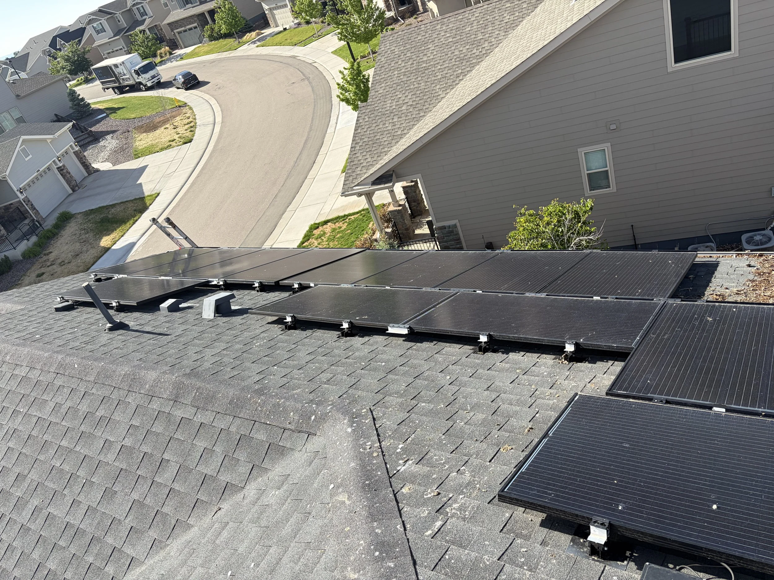 View of a neighborhood street from a rooftop with solar panels. Houses with driveways, trees, and a parked truck and car are visible. The street curves around the neighborhood.
