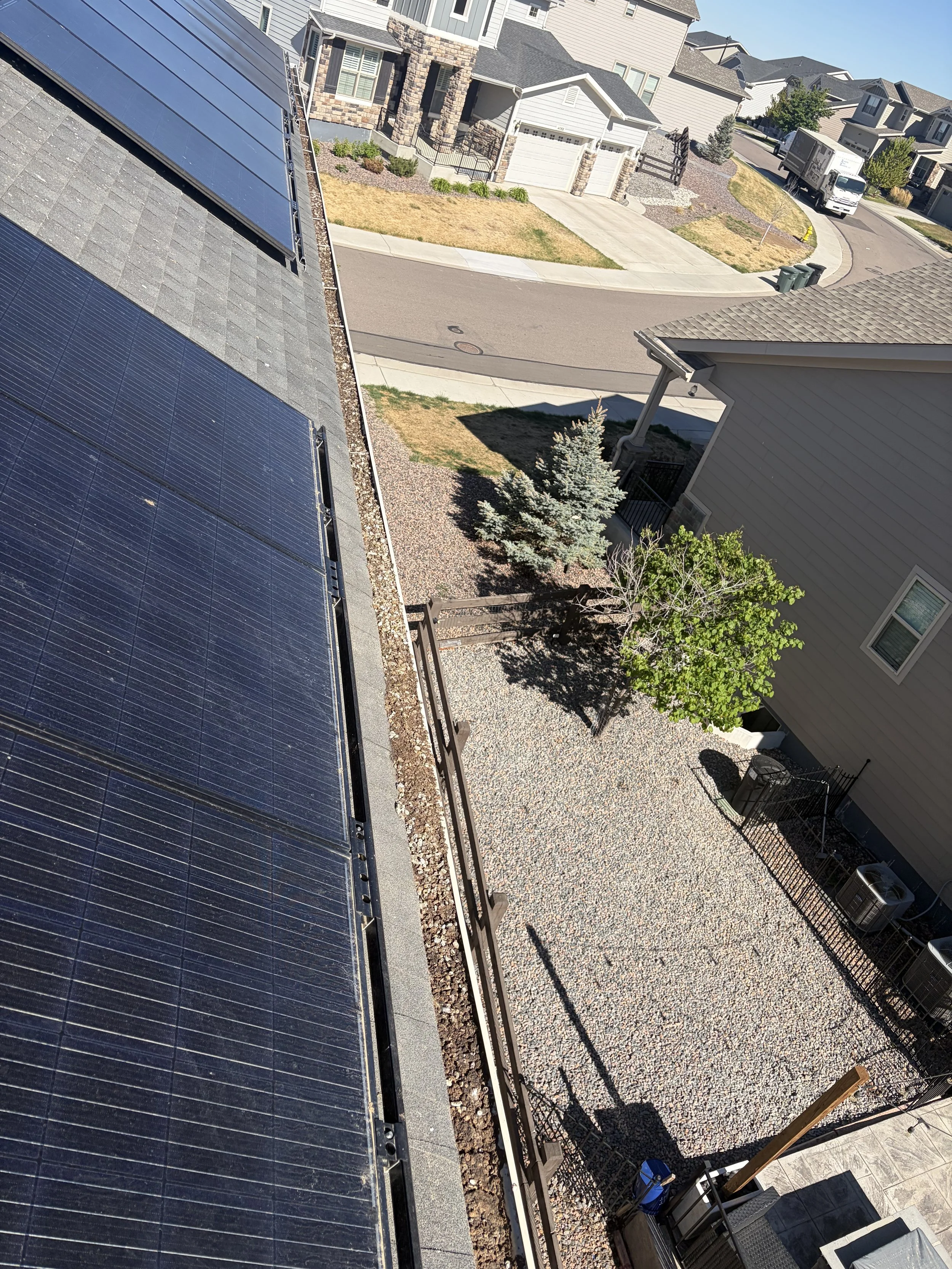 Residential neighborhood with houses, a curved street, and a parked truck. The image is taken from an elevated position showing solar panels installed on the roof in the foreground.