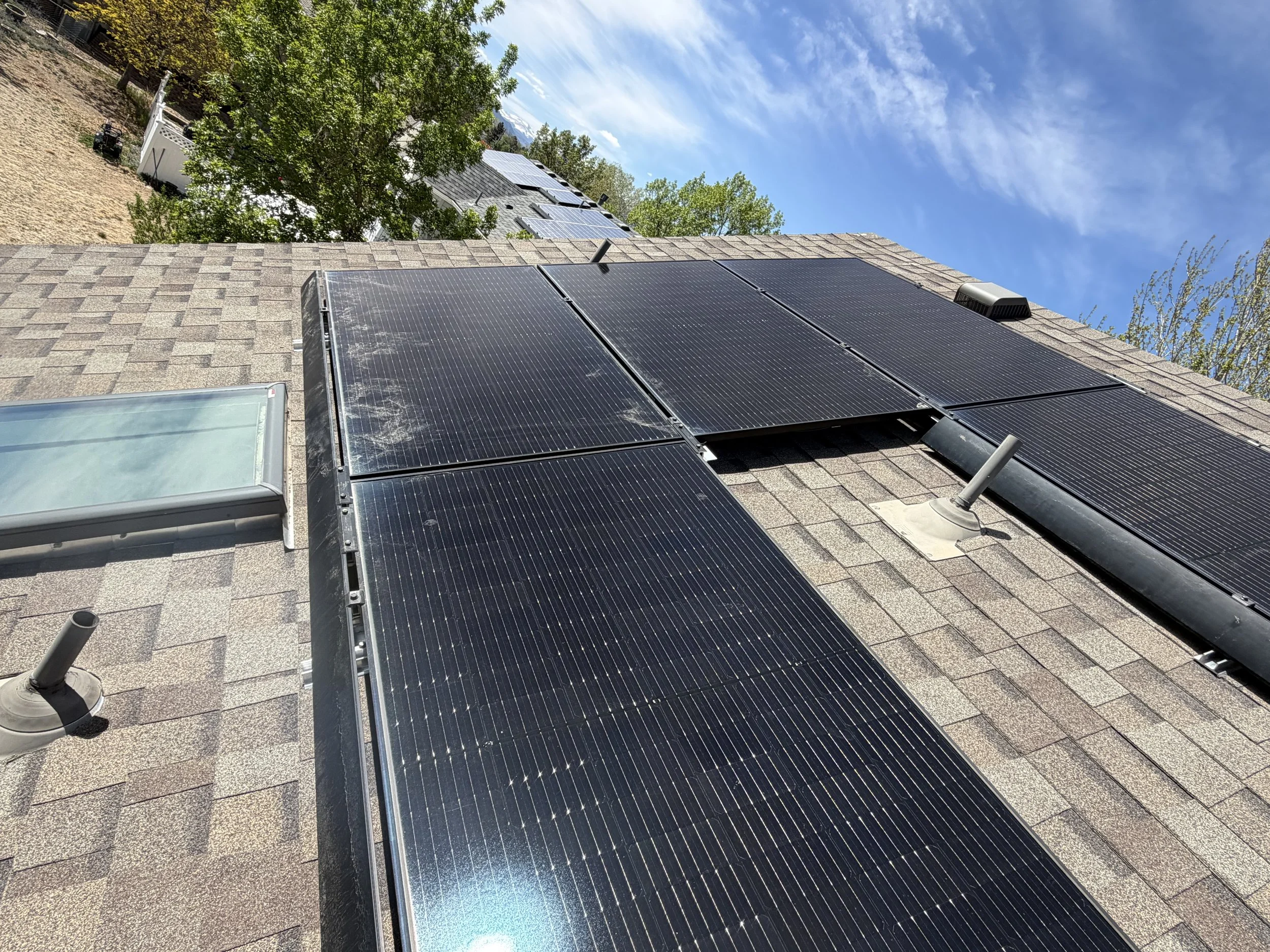 The image shows a house roof with black solar panels installed. There are trees in the background and a partly cloudy sky.