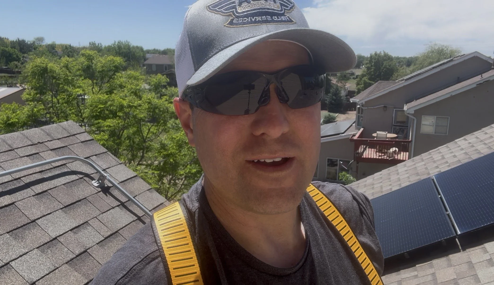 A man wearing a gray cap, sunglasses, and a gray shirt with a yellow strap. He is on a rooftop with solar panels and solar hot water heaters, with residential houses, trees, and a partly cloudy sky in the background.