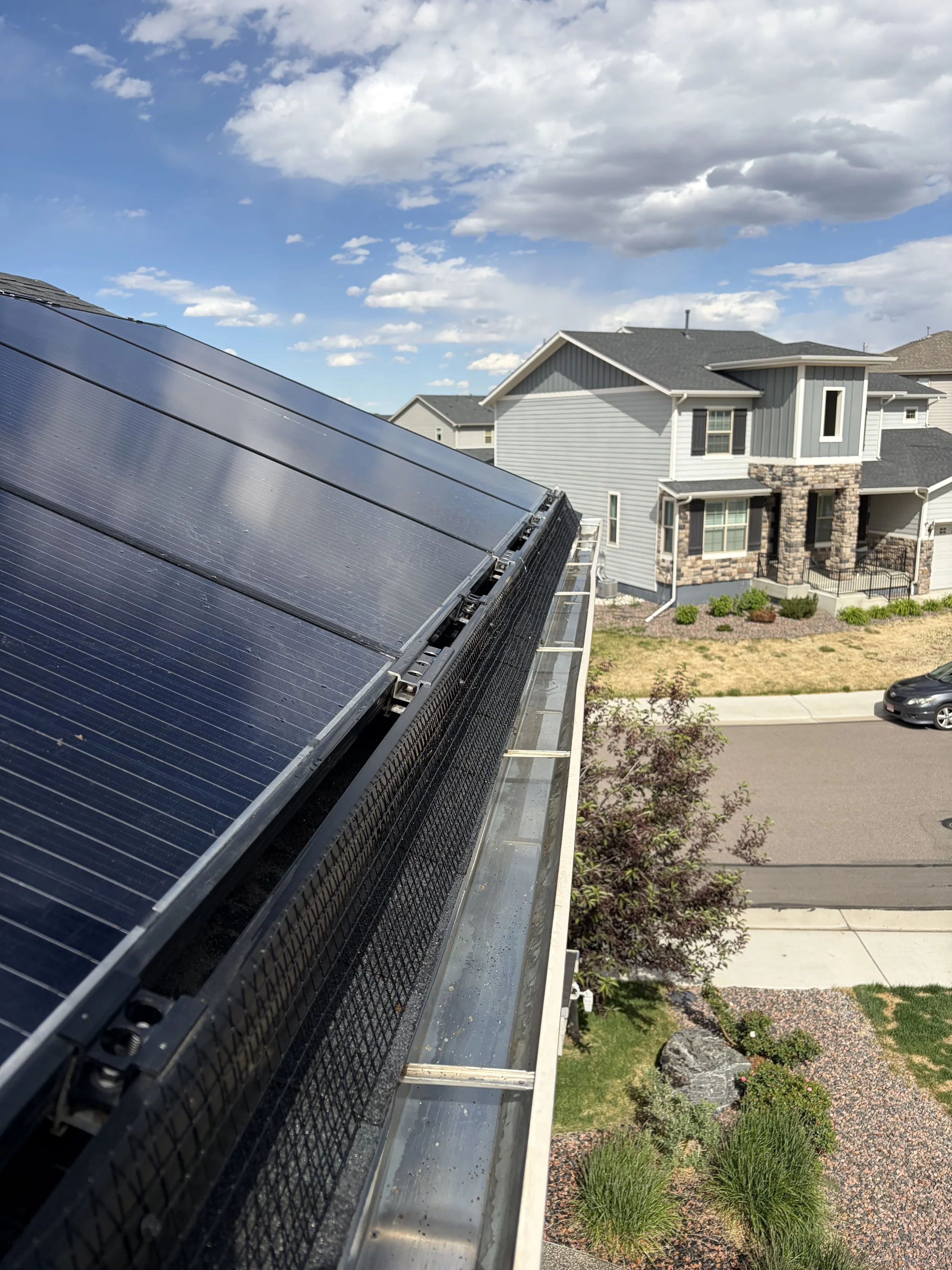Photograph of solar panels installed on the roof of a residential house under a partly cloudy sky, with other houses, a street, a car, and landscaped yards visible in the background.
