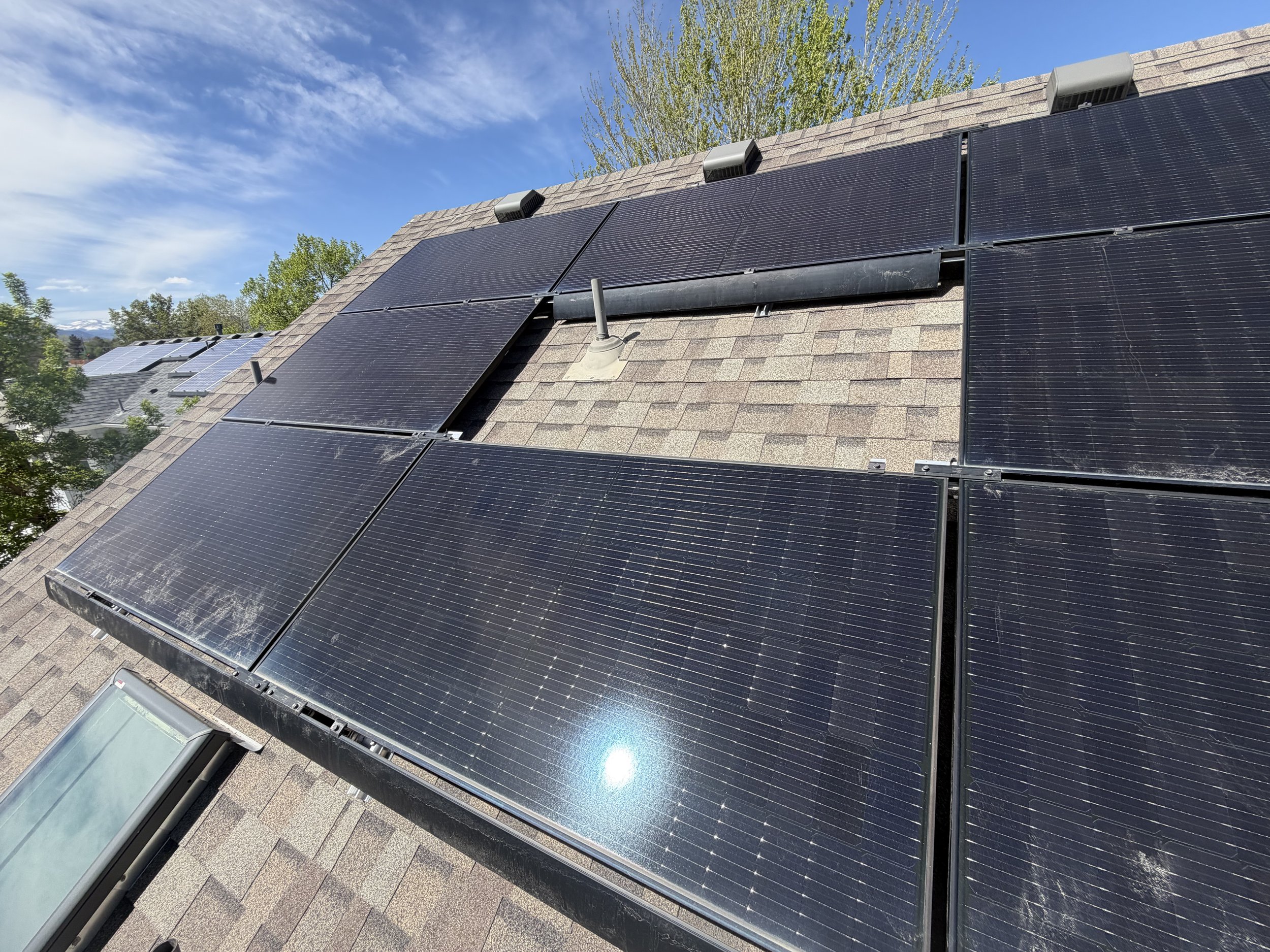 Several black solar panels installed on a sloped roof under a partly cloudy blue sky, with green trees in the background.