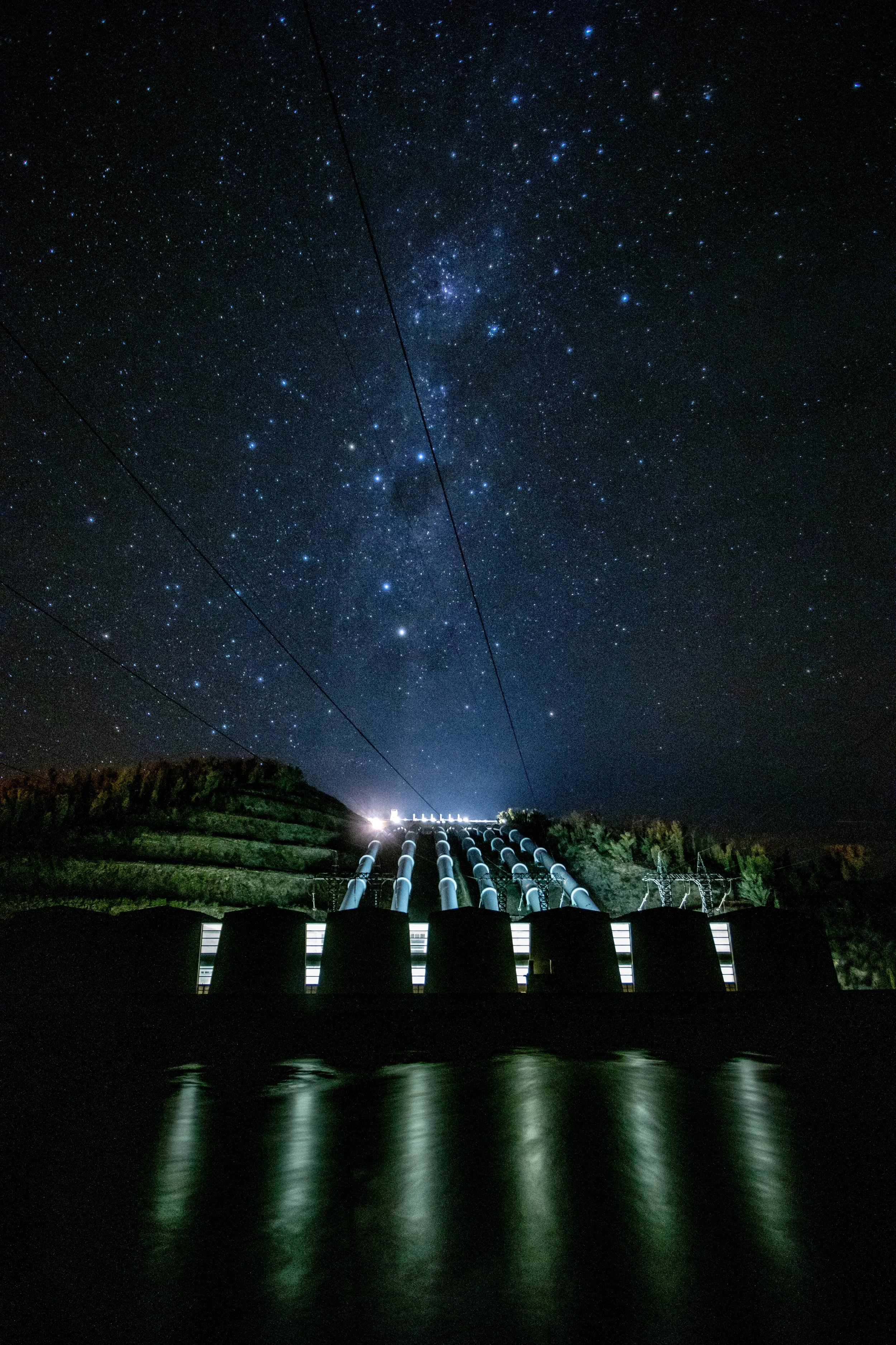 Nighttime view of a hydroelectric dam with water reflecting lights, under a starry sky with visible stars and the Milky Way galaxy.
