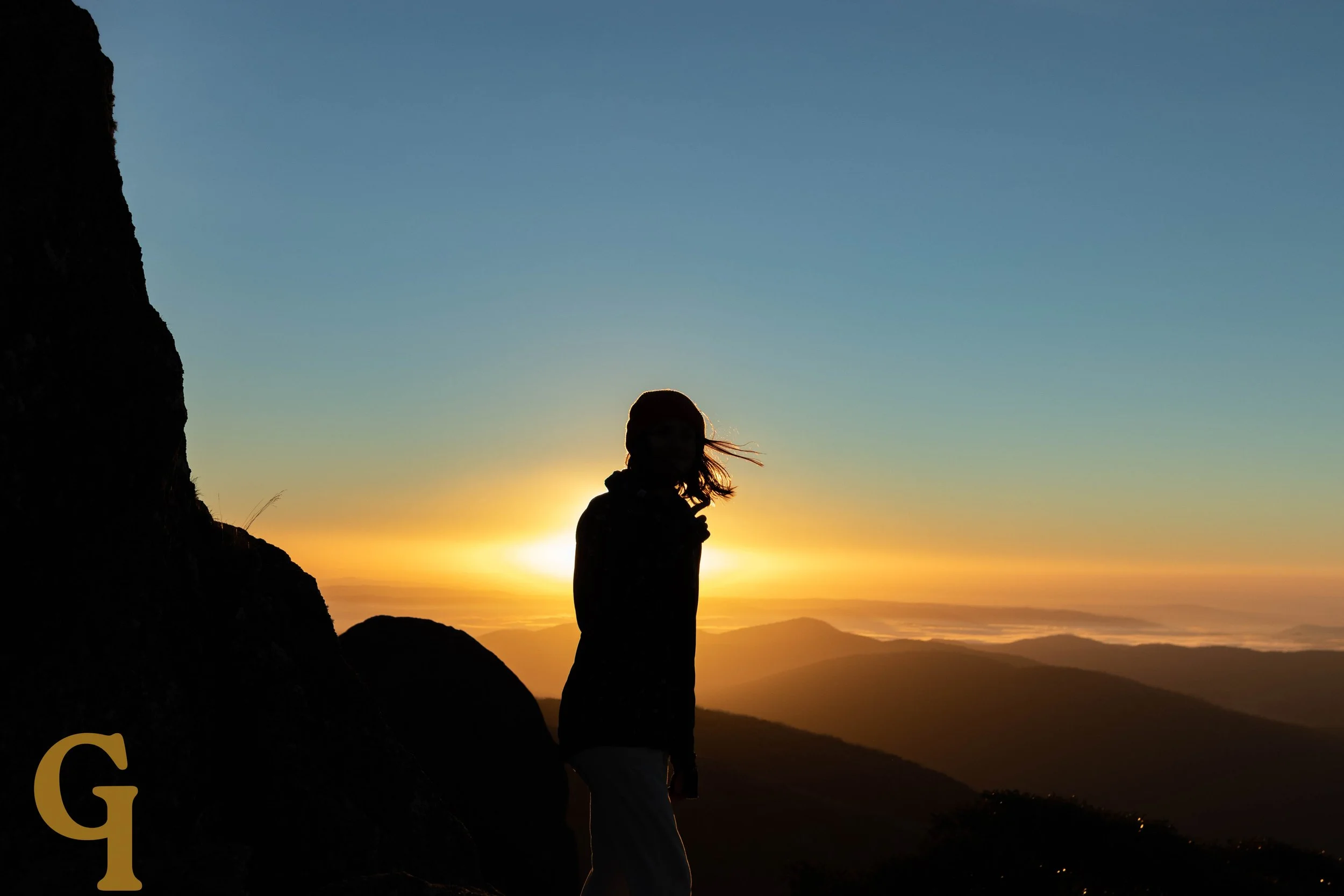 A person standing on a mountain at sunset with a backdrop of mountains and a colorful sky.