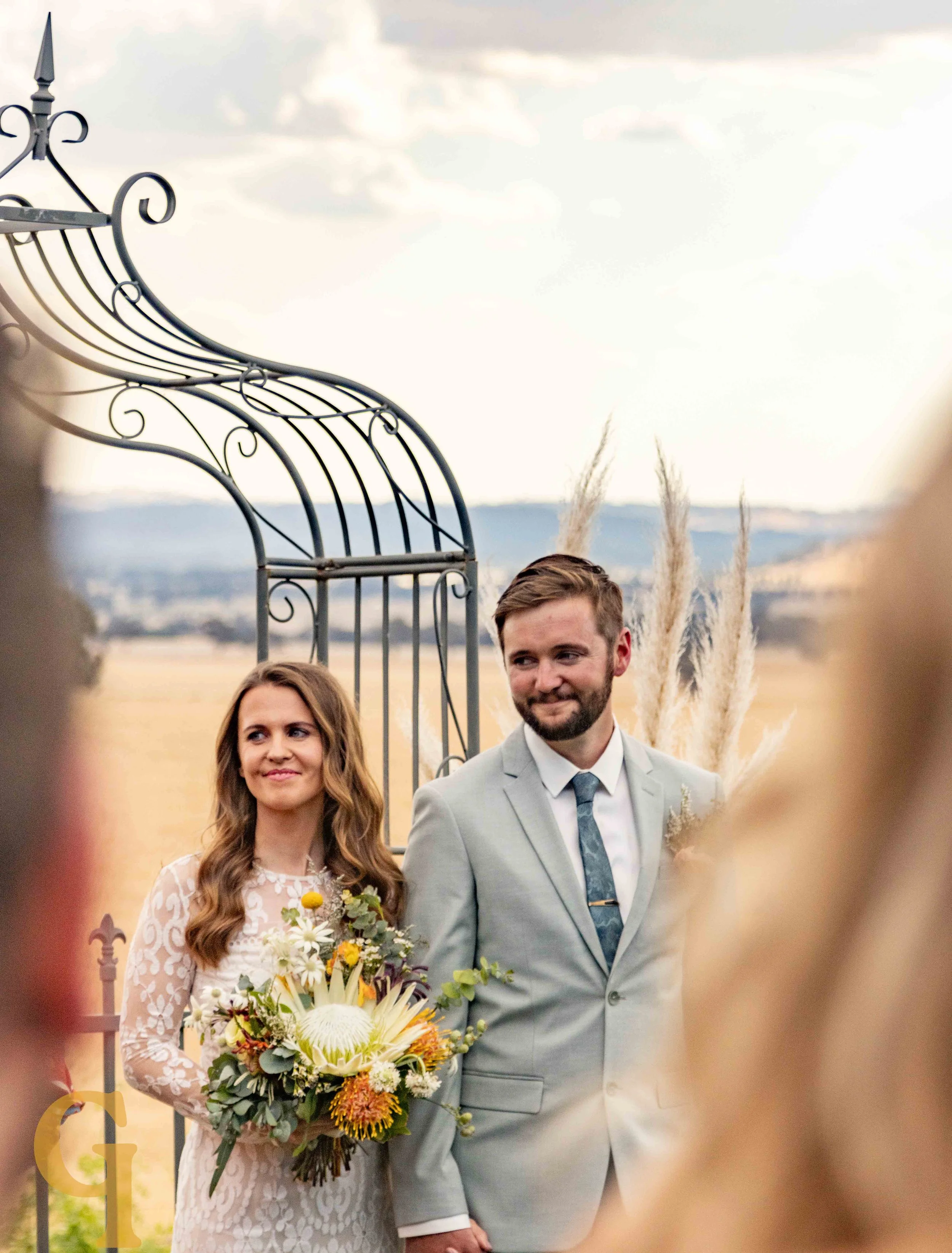Bride holding a bouquet standing next to groom in a gray suit during outdoor wedding ceremony.
