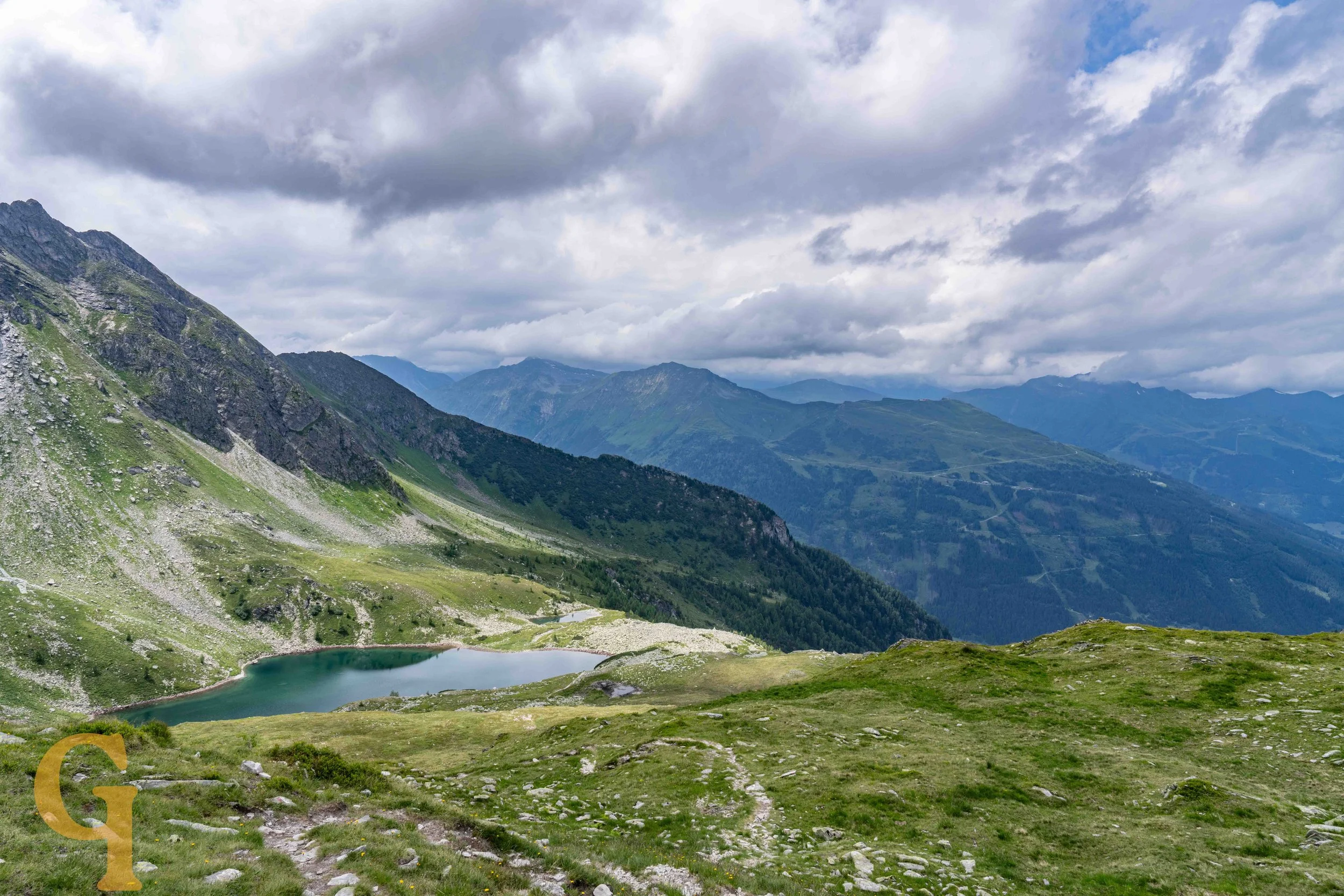 Mountain landscape with a small lake in the foreground, green rolling hills, and tall mountains under a cloudy sky.