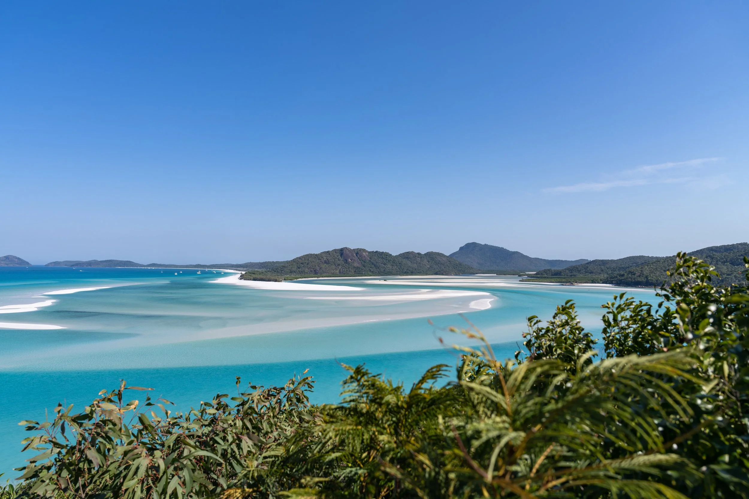 A scenic view of a beach with white sandbars, turquoise water, green islands in the background, and clear blue sky.