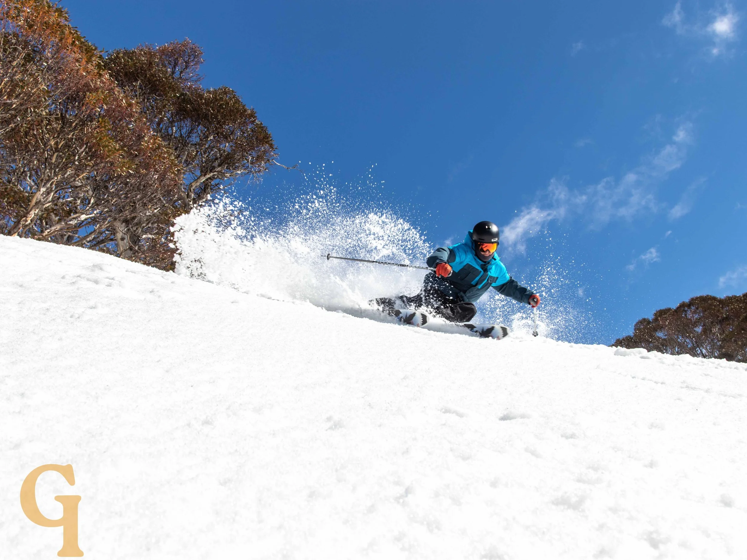 A skier wearing a blue jacket, black helmet, and orange goggles skiing down a snowy slope under a clear blue sky with trees in the background.