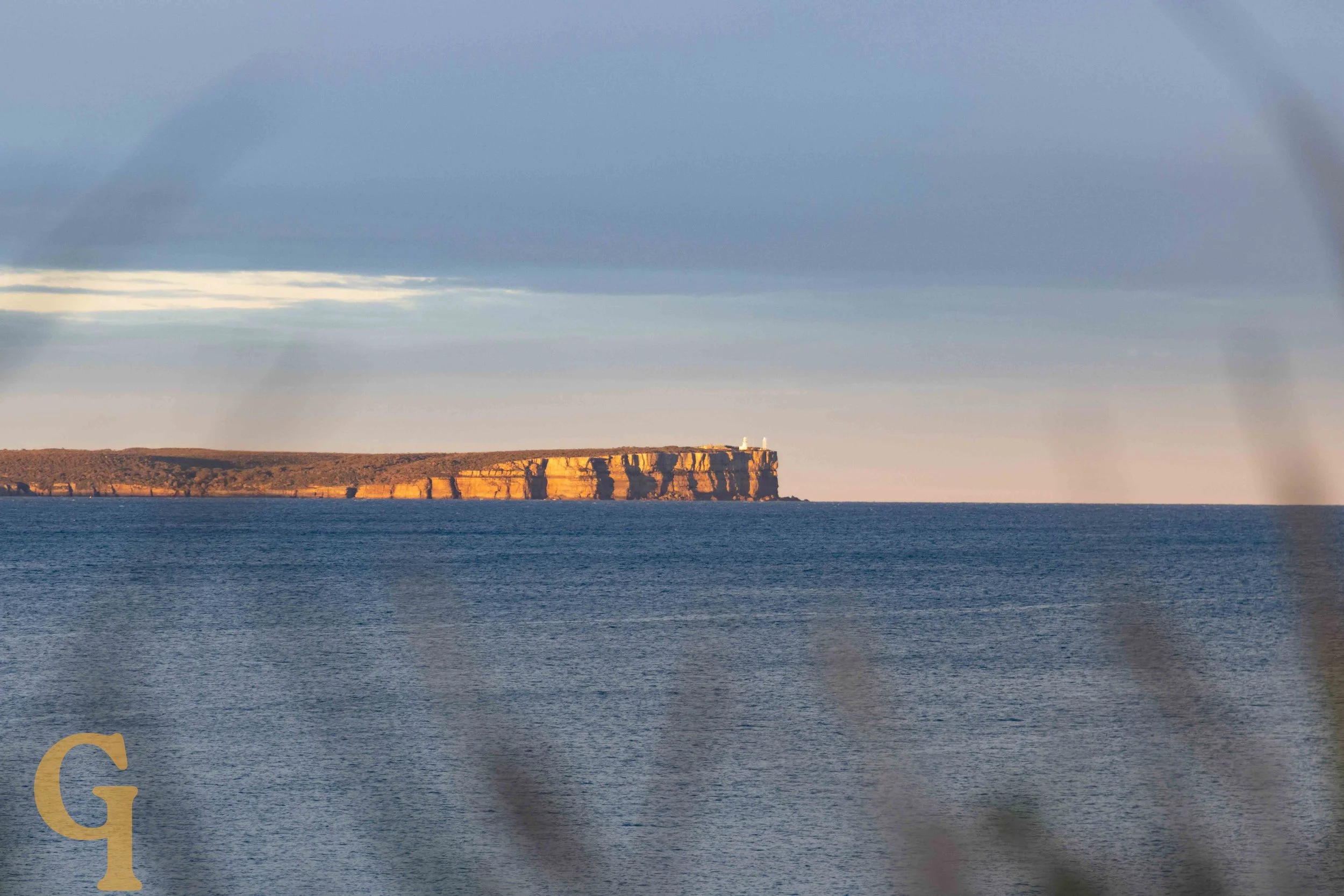 View of a distant cliffside with a lighthouse on top, seen across a body of water under a cloudy sky.
