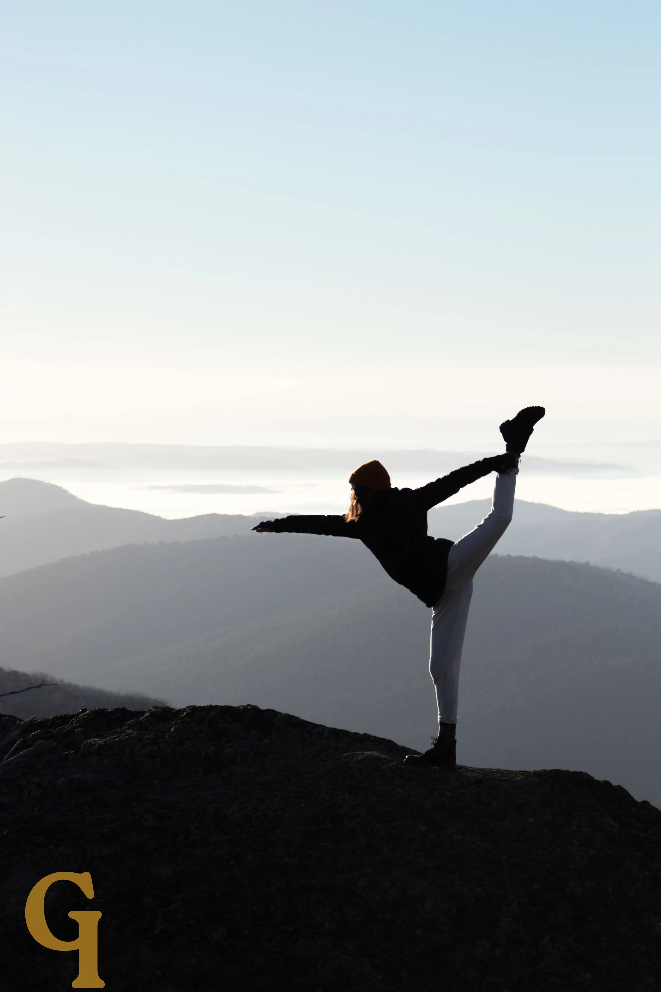 Person performing a yoga pose on mountain summit during sunset or sunrise, with scenic mountain landscape in background.