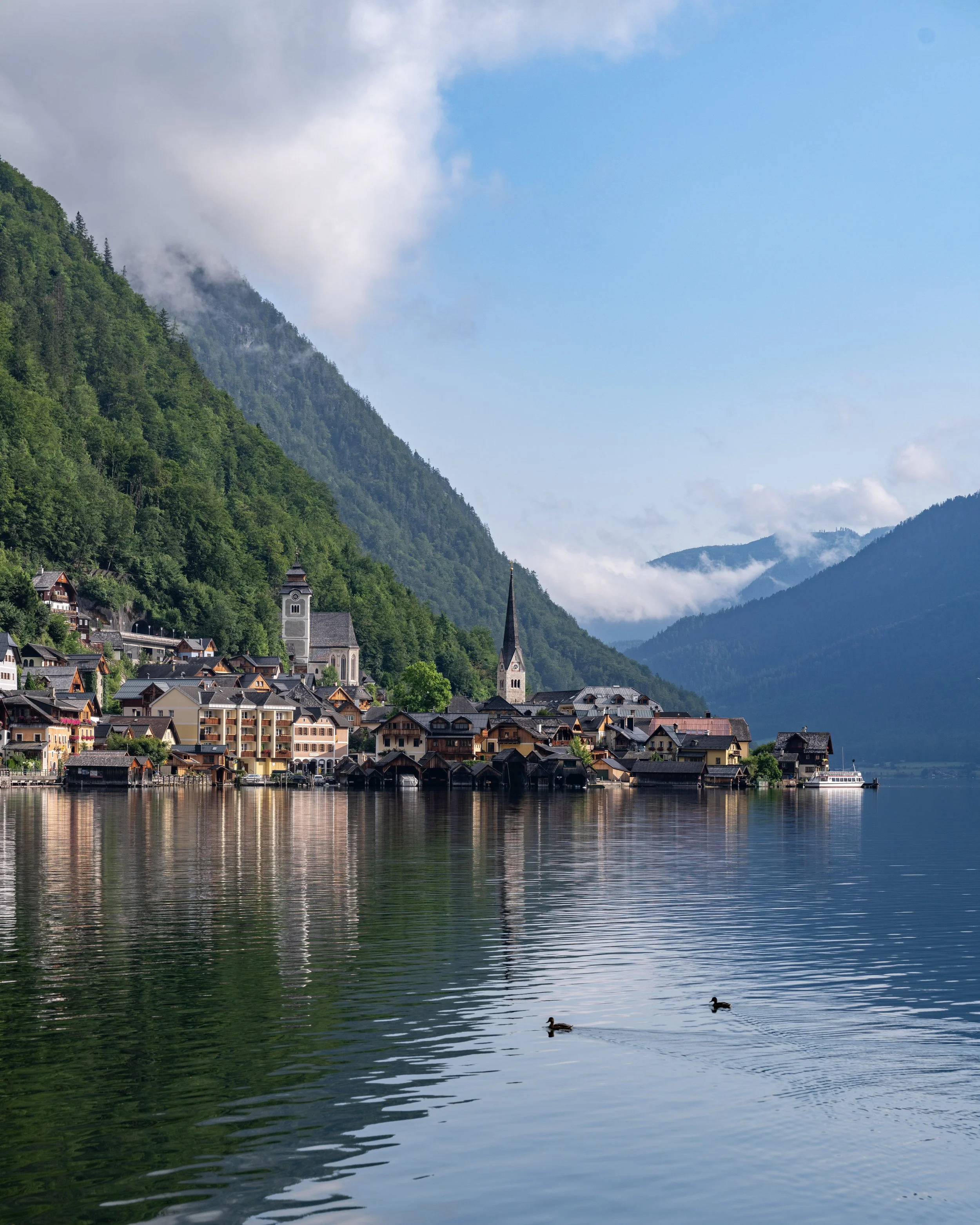 A scenic view of a lakeside town with colorful houses, a church steeple, and lush green mountains in the background, with ducks swimming in the calm water.