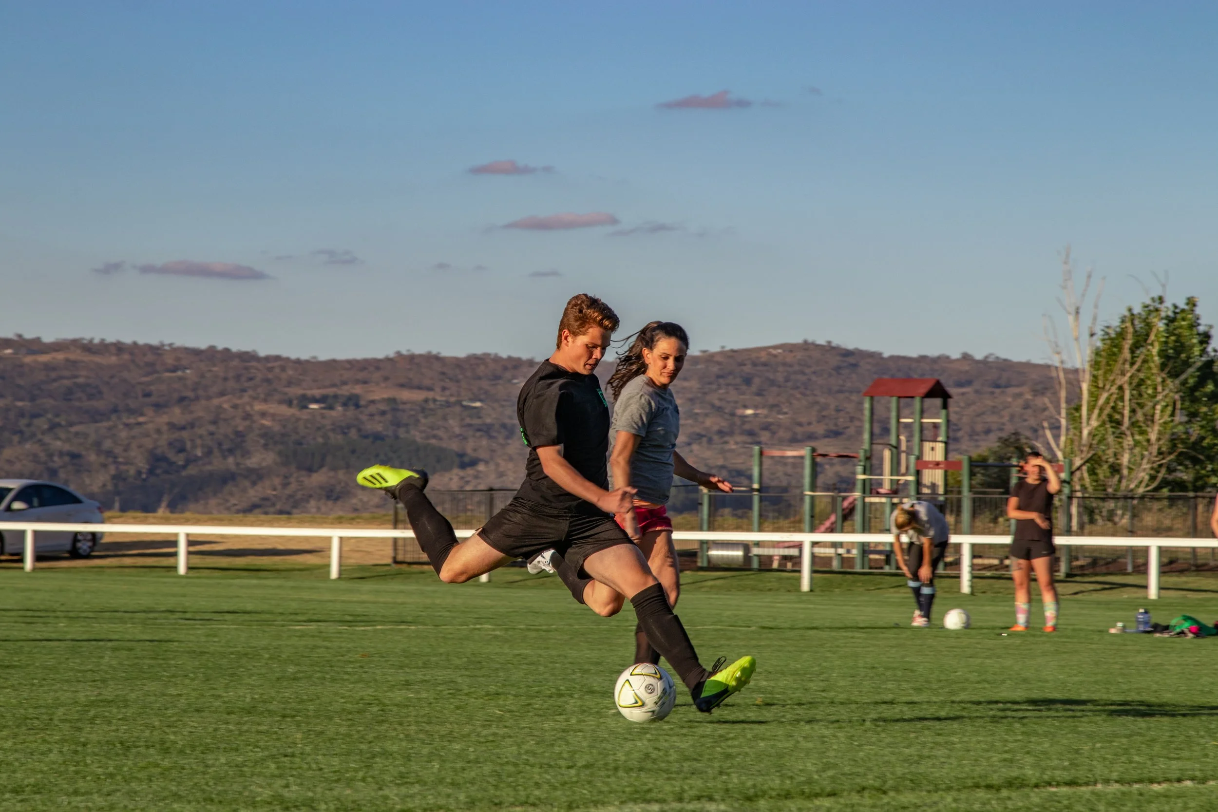 Two female soccer players in black and gray shirts compete for the ball on a grass field, with a playground, trees, and mountains in the background.