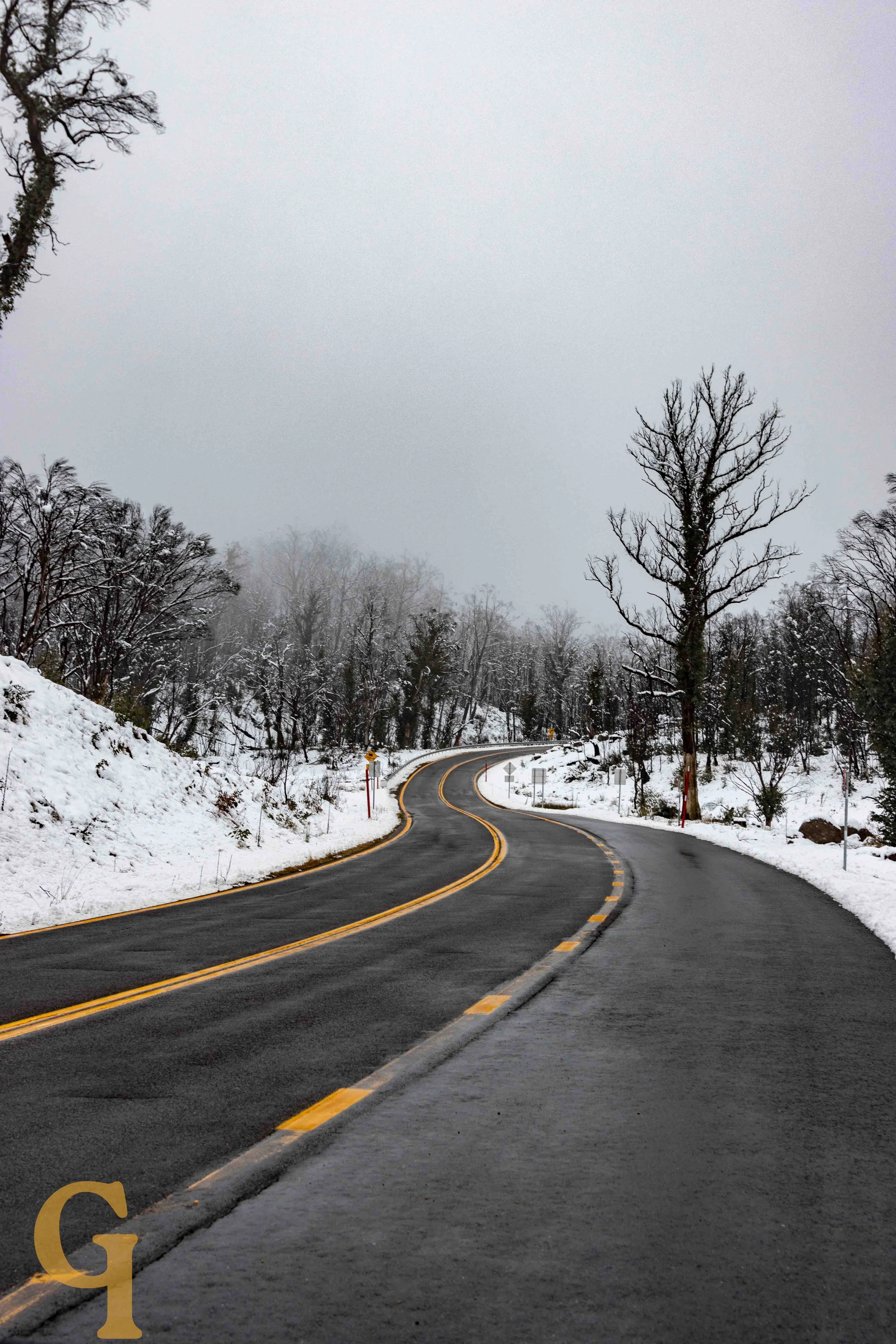 Winding mountain road with snow on sides and leafless trees, overcast sky.