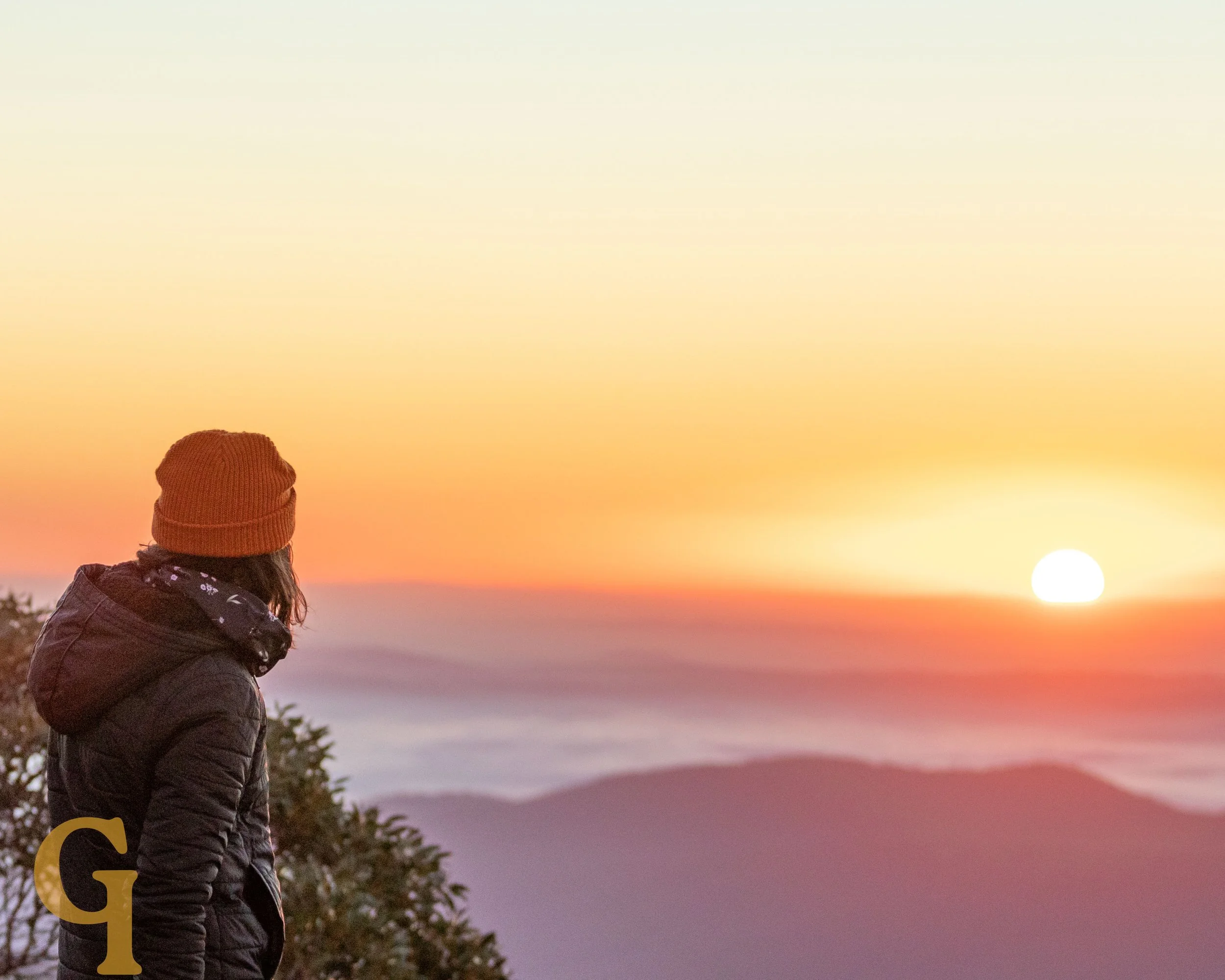 Person wearing a brown beanie and a black jacket looking at a sunrise over mountains.