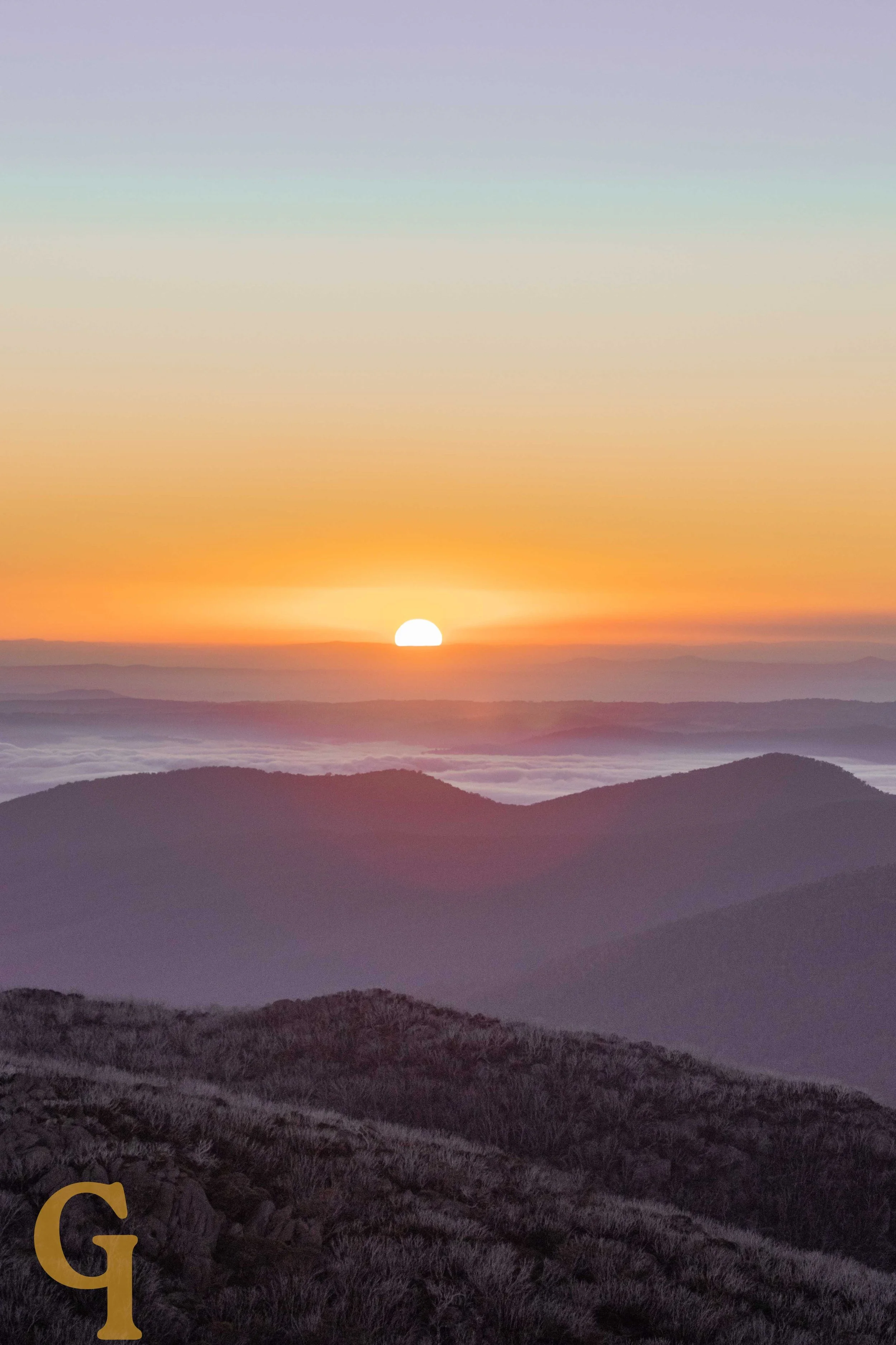 Sunset over mountain range with a colorful sky filled with orange, pink, and purple hues, and layers of mist or clouds in the valleys between the hills.