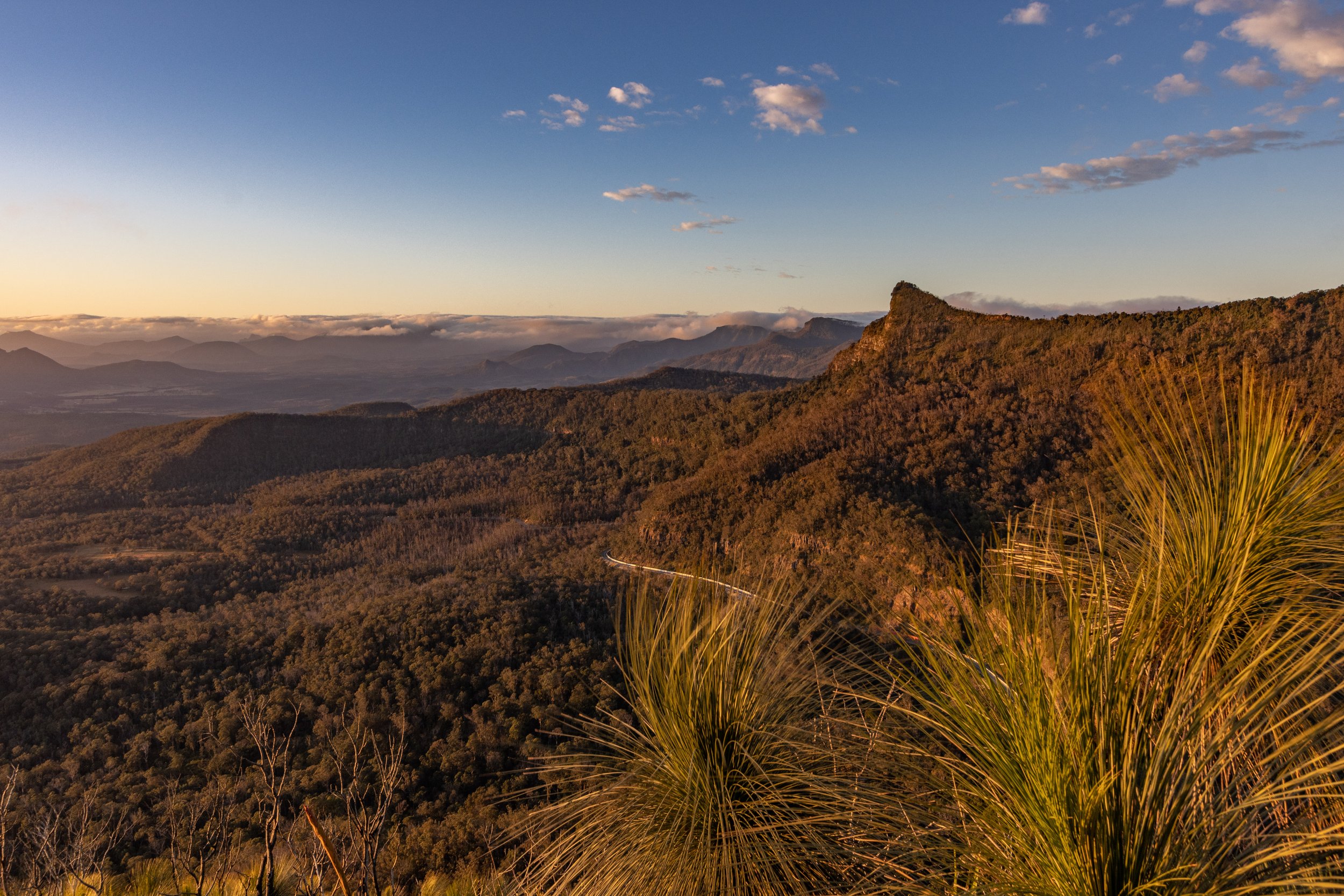 Sunset over a mountain range with a winding road visible near the bottom and a clear sky with some clouds.