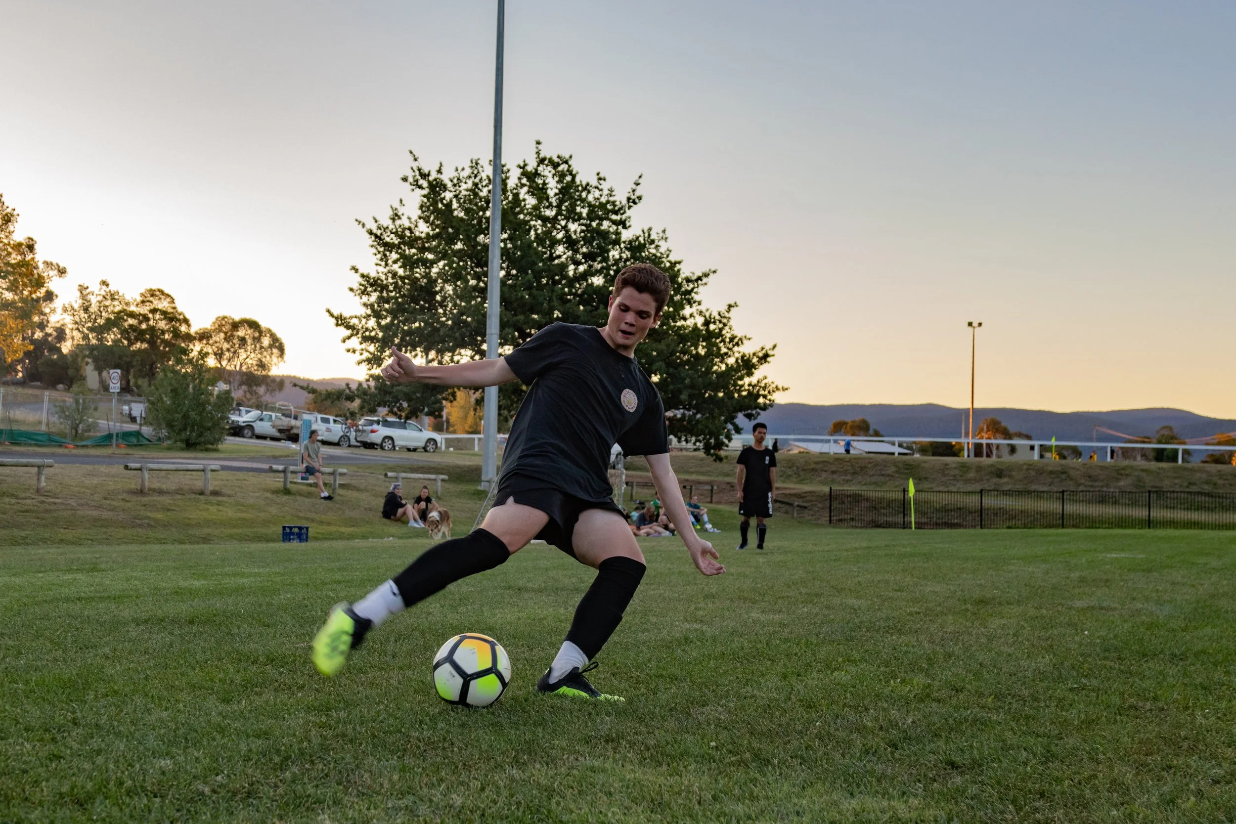 A young male soccer player in black attire and bright yellow shoes kicks a soccer ball on a grassy field during sunset, with people and cars in the background.