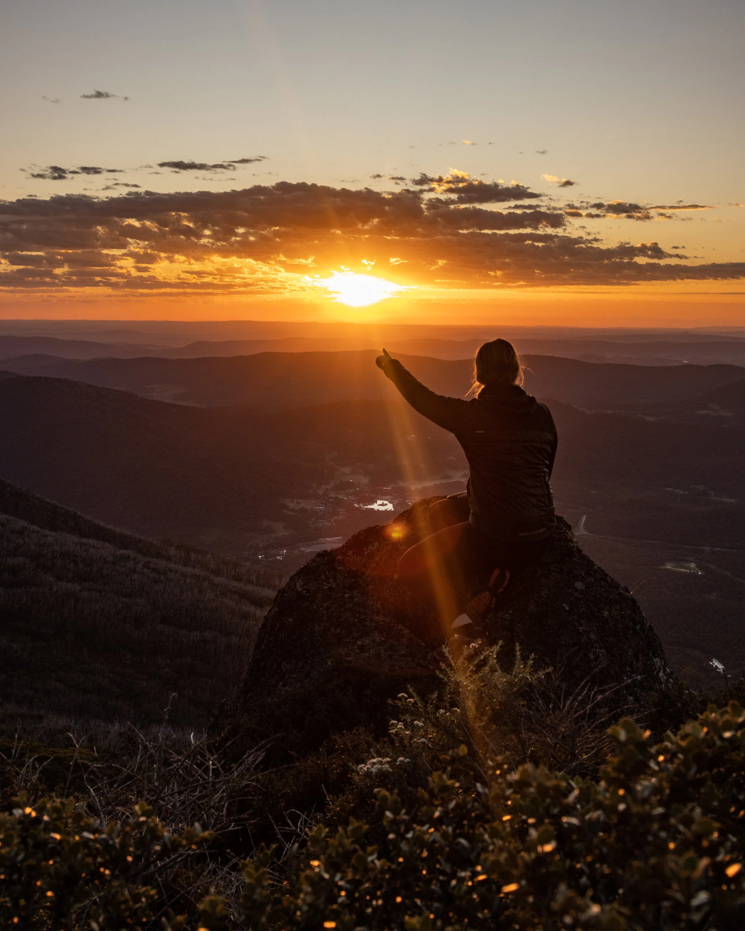 Person sitting on a rock at sunset, pointing at the sky over a mountainous landscape.