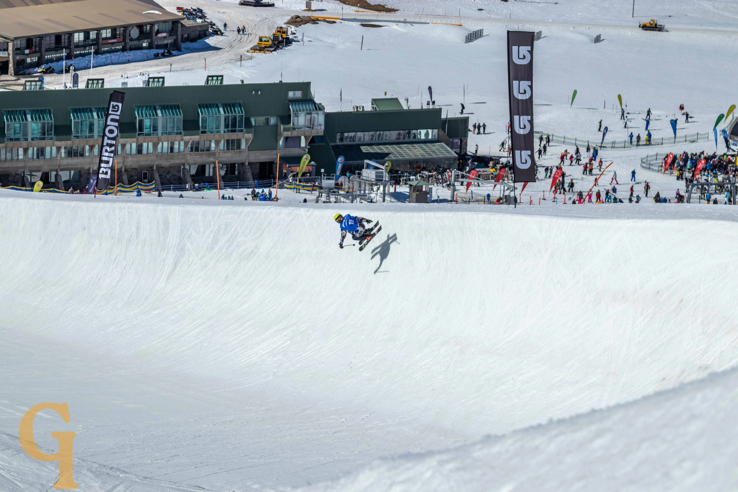 A snowboarder mid-air performing a trick on a snowy halfpipe at a ski resort.