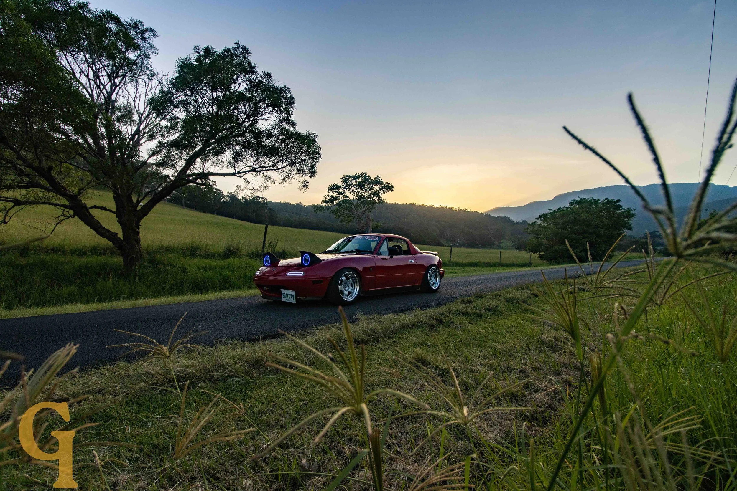 A red Mazda MX-5 Miata with cat ears and glowing blue eyes headlights, parked on a country road at sunset, with trees and hills in the background.