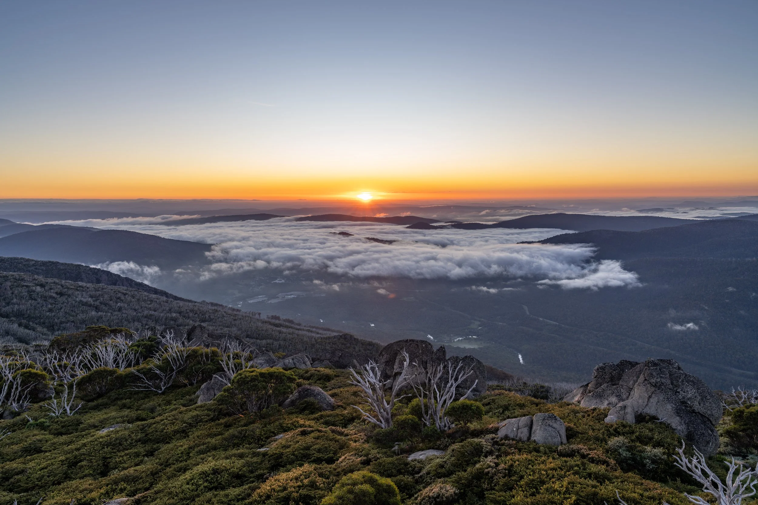 Sunrise over a mountain landscape with mist and clouds, rocky foreground with sparse trees and shrubs.