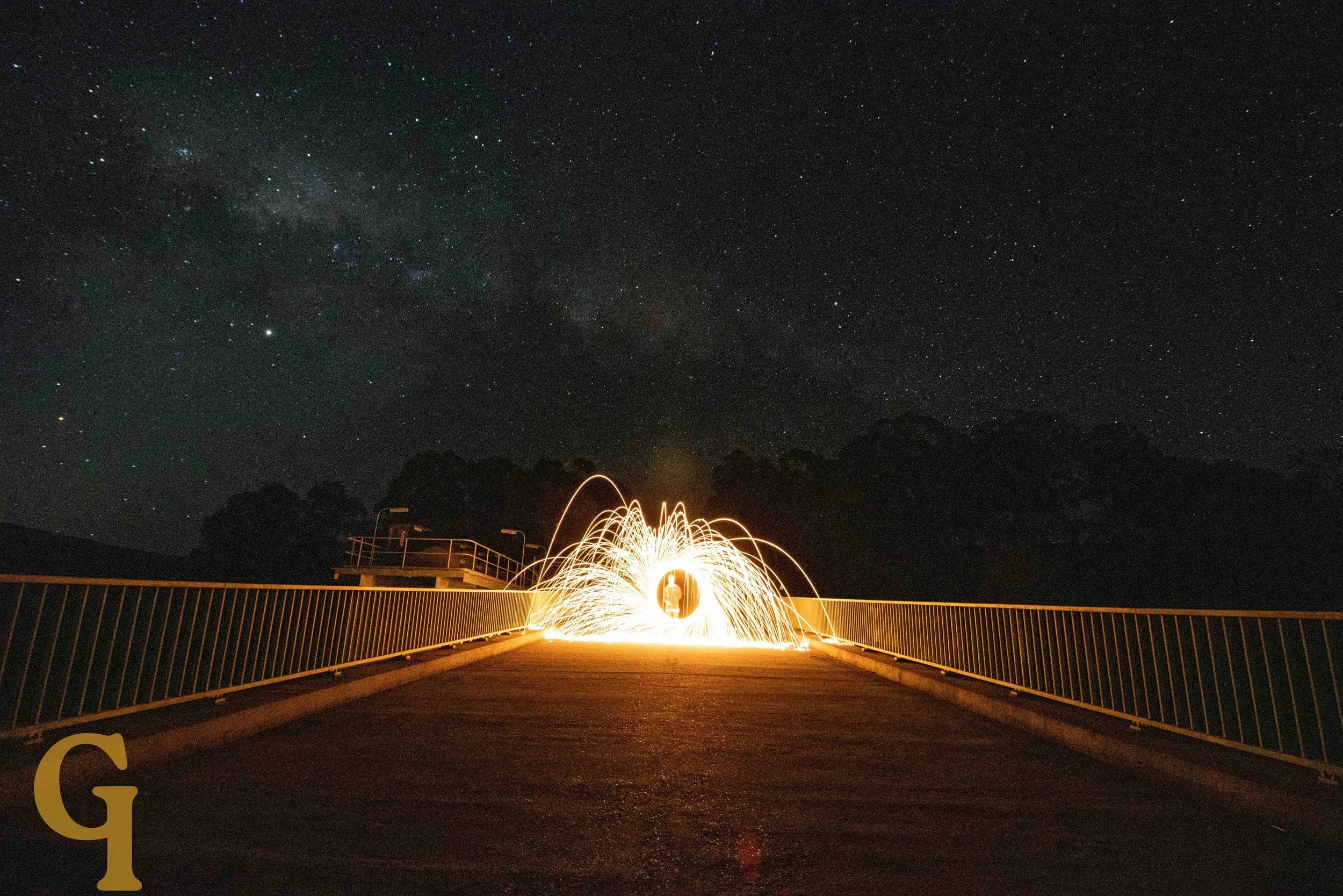 Person creating sparks with steel wool on a bridge at night, under a starry sky with the Milky Way visible.