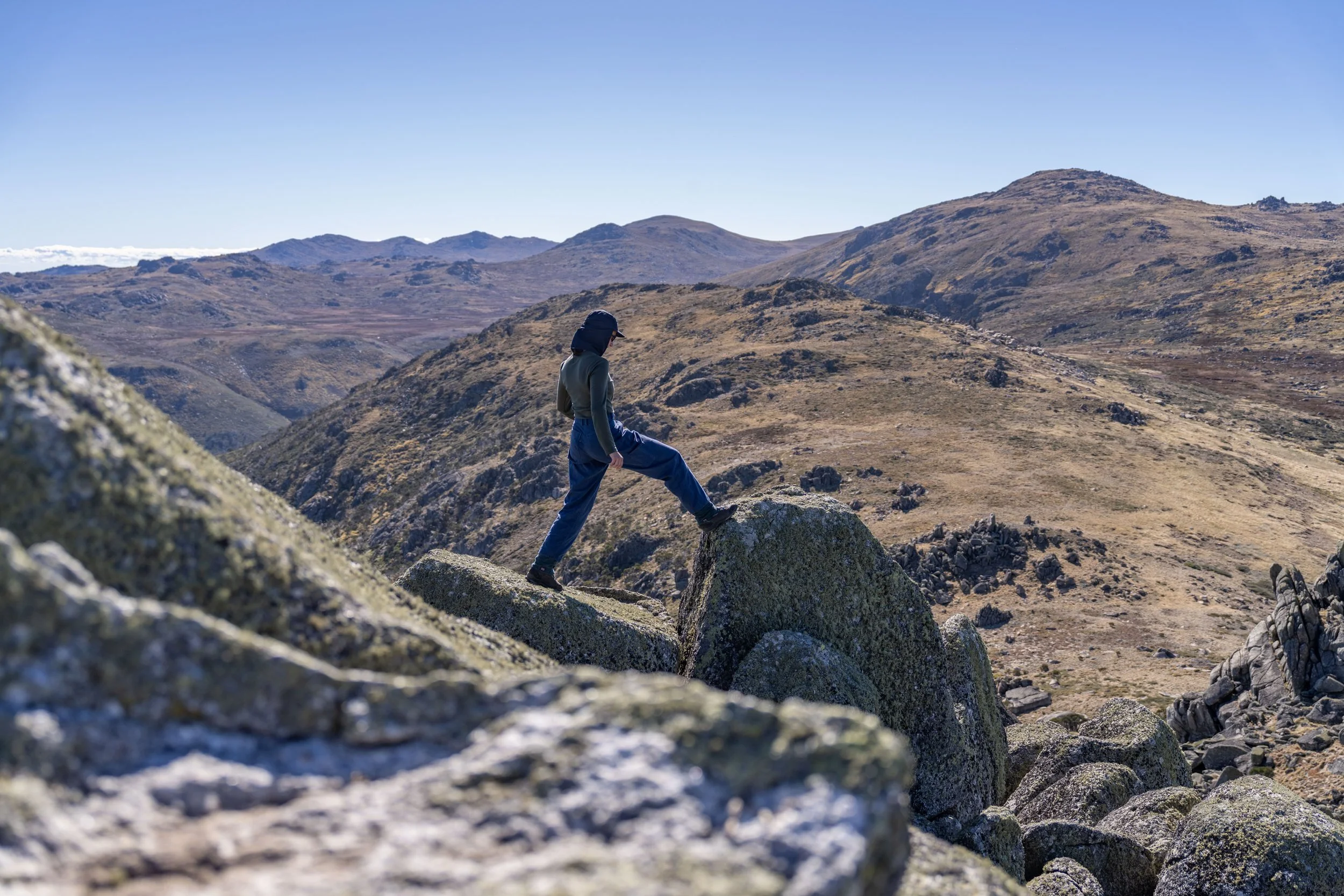 Person wearing a hat and outdoor clothing hiking on large rocks in the Snowy Mountains, NSW with rolling hills and clear blue sky.