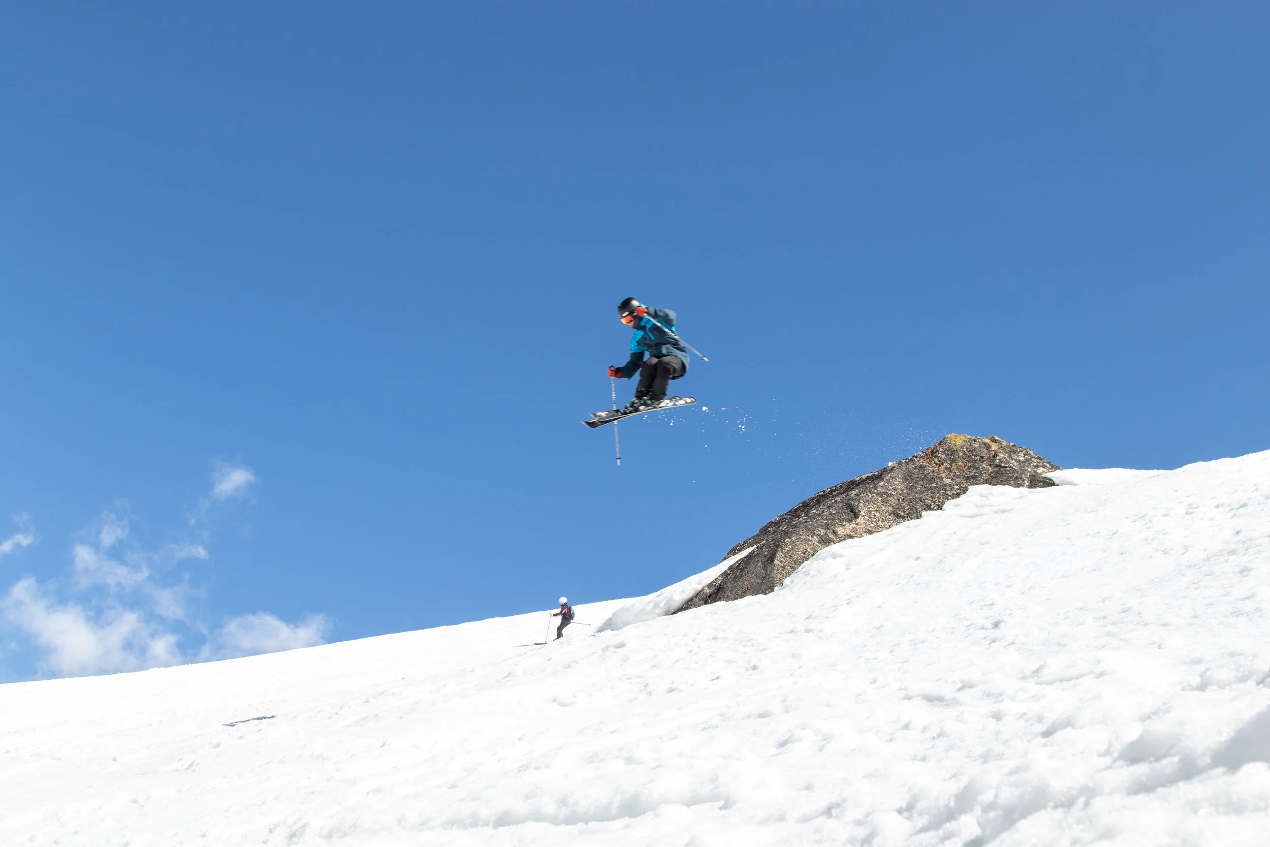 A skier in a blue jacket and black pants jumping off a snow-covered slope with a clear blue sky in the background.
