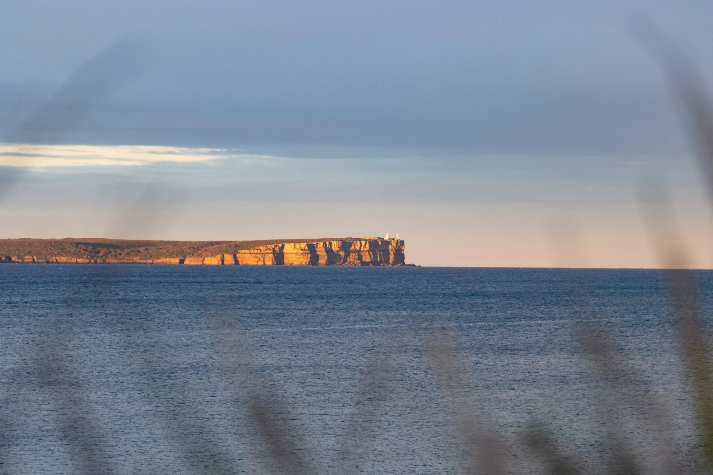View of a distant cliffside with a lighthouse on top, seen across a body of water under a cloudy sky.