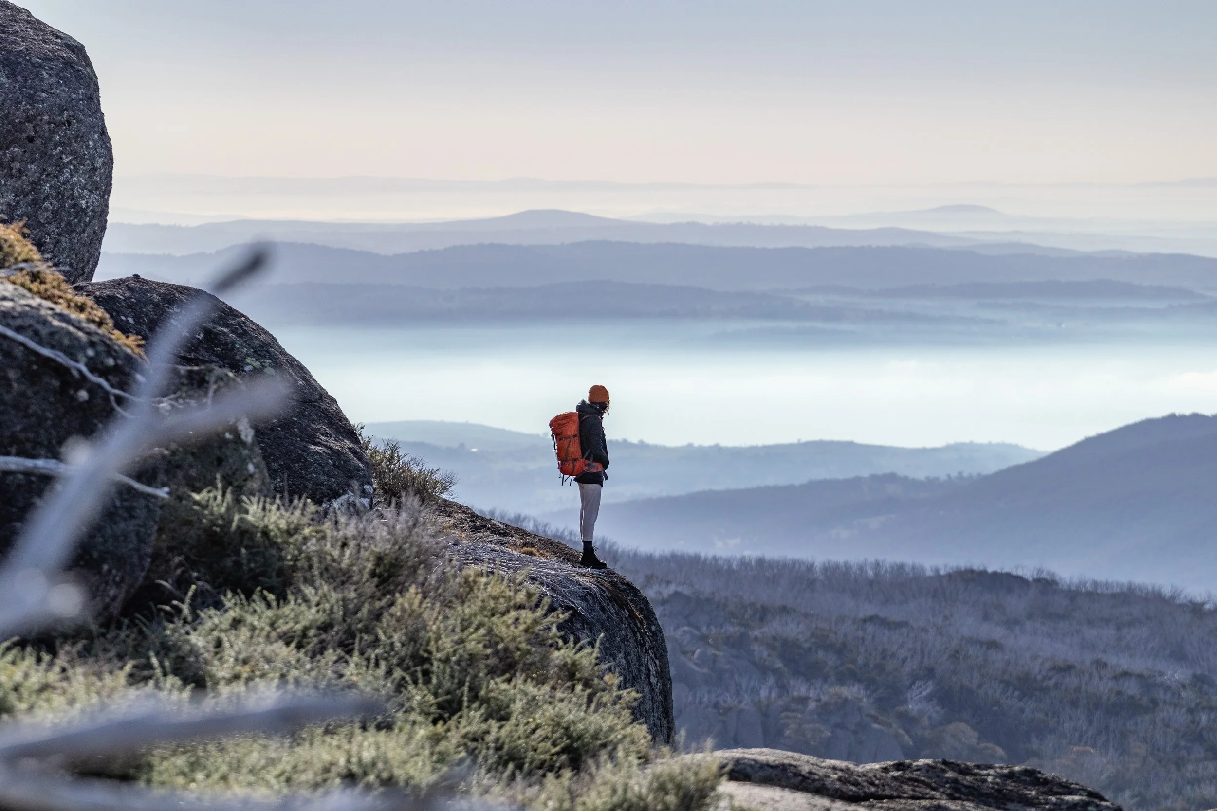 A hiker with a backpack standing on a rocky mountain ledge overlooking a misty, layered landscape of hills and water bodies.
