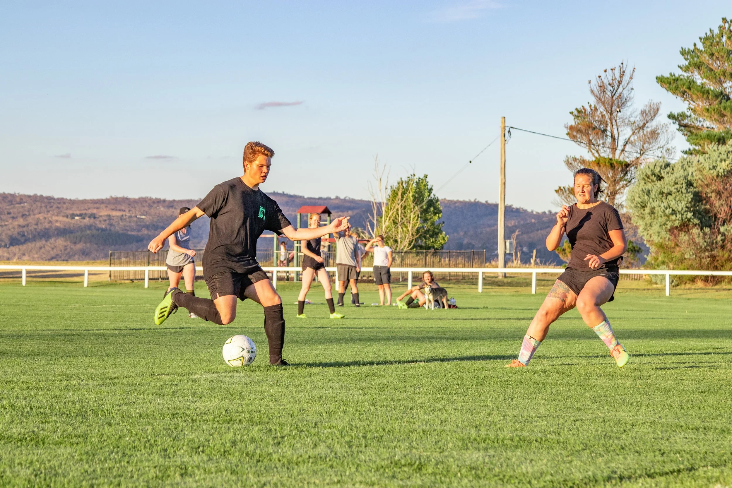 A group of people playing soccer on a grassy field, with two women actively competing for the ball, and others watching or relaxing in the background under clear skies.