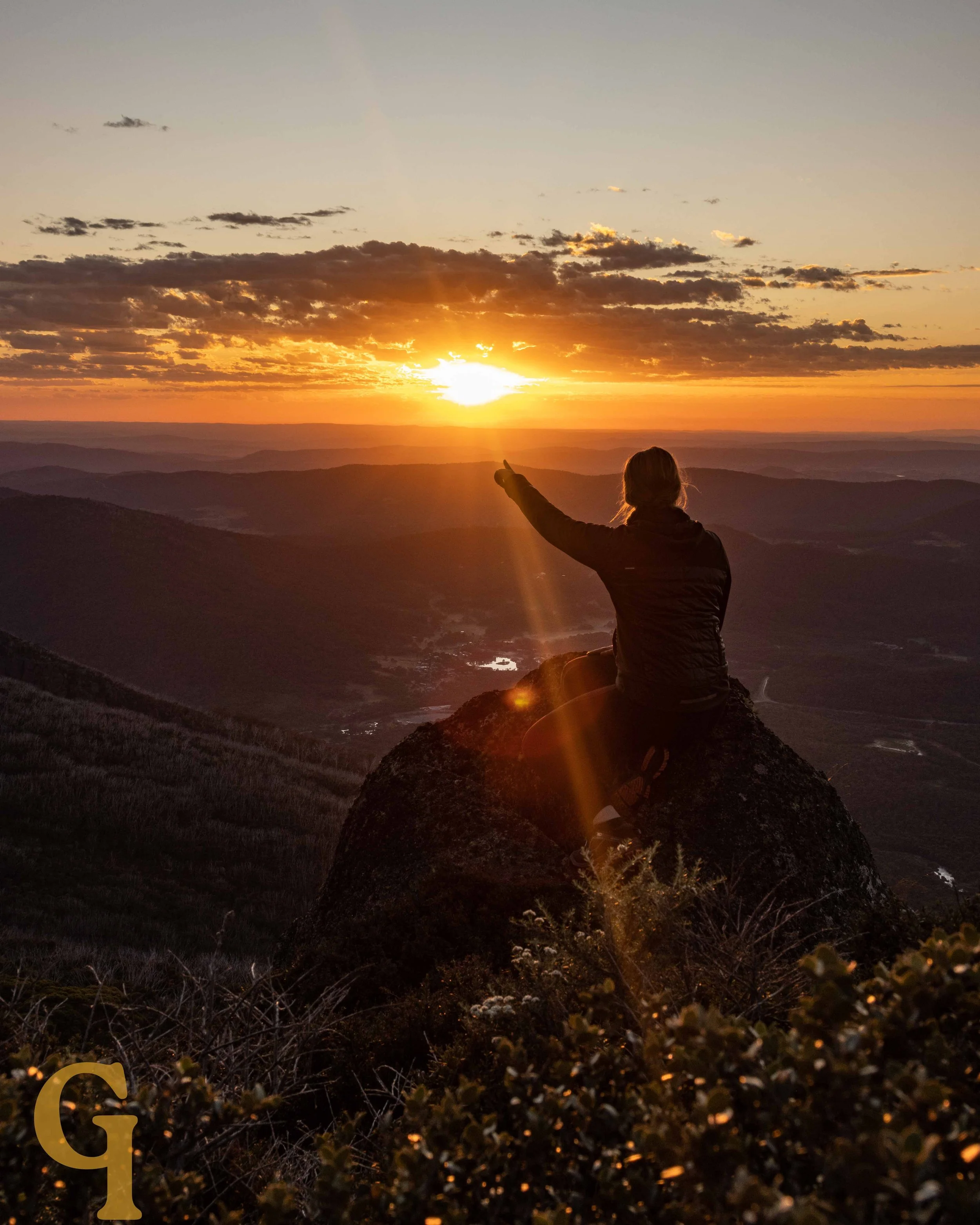 Person sitting on a rock at sunset, pointing at the sky over a mountainous landscape.