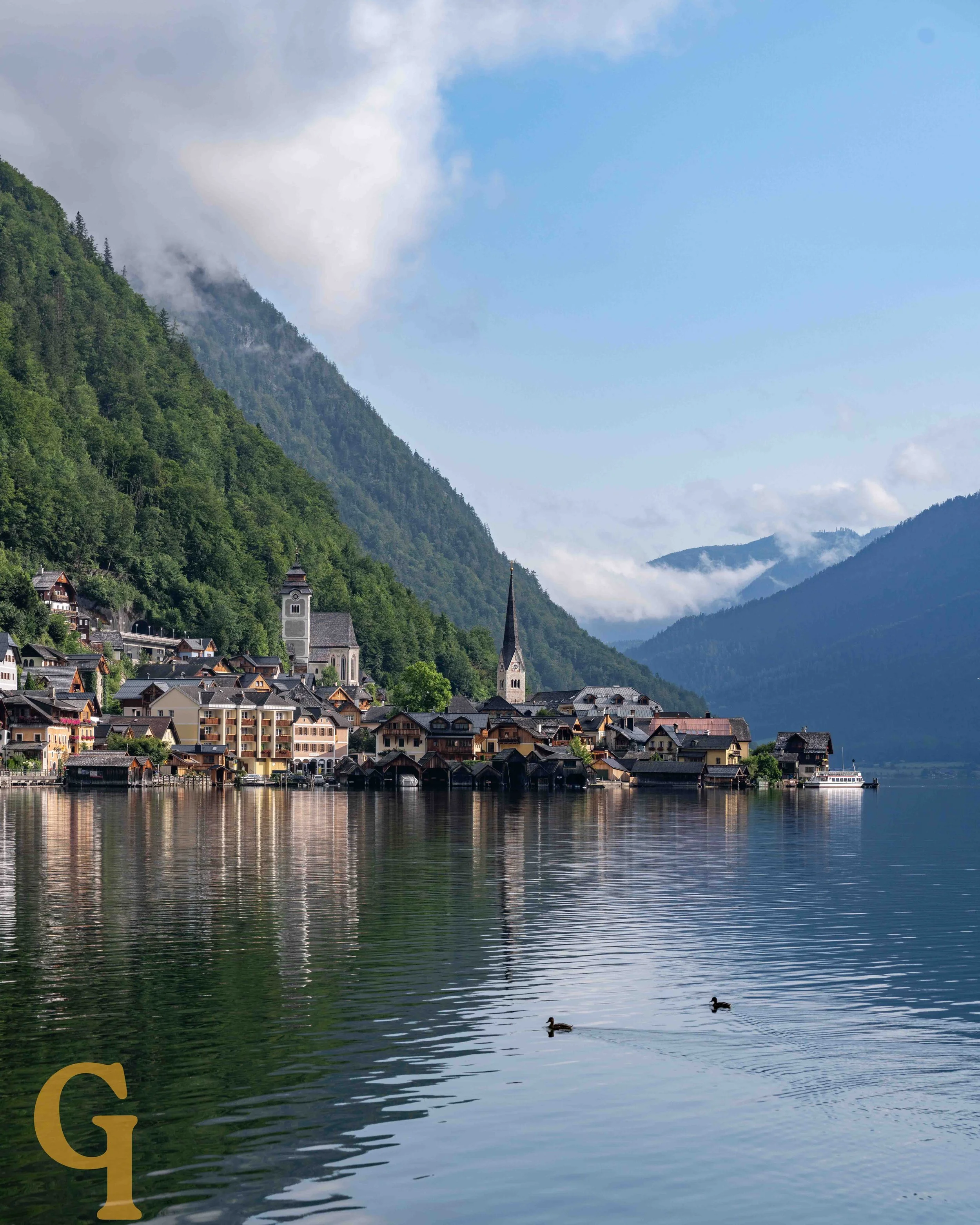 A scenic view of a lakeside town with colorful houses, a church steeple, and lush green mountains in the background, with ducks swimming in the calm water.