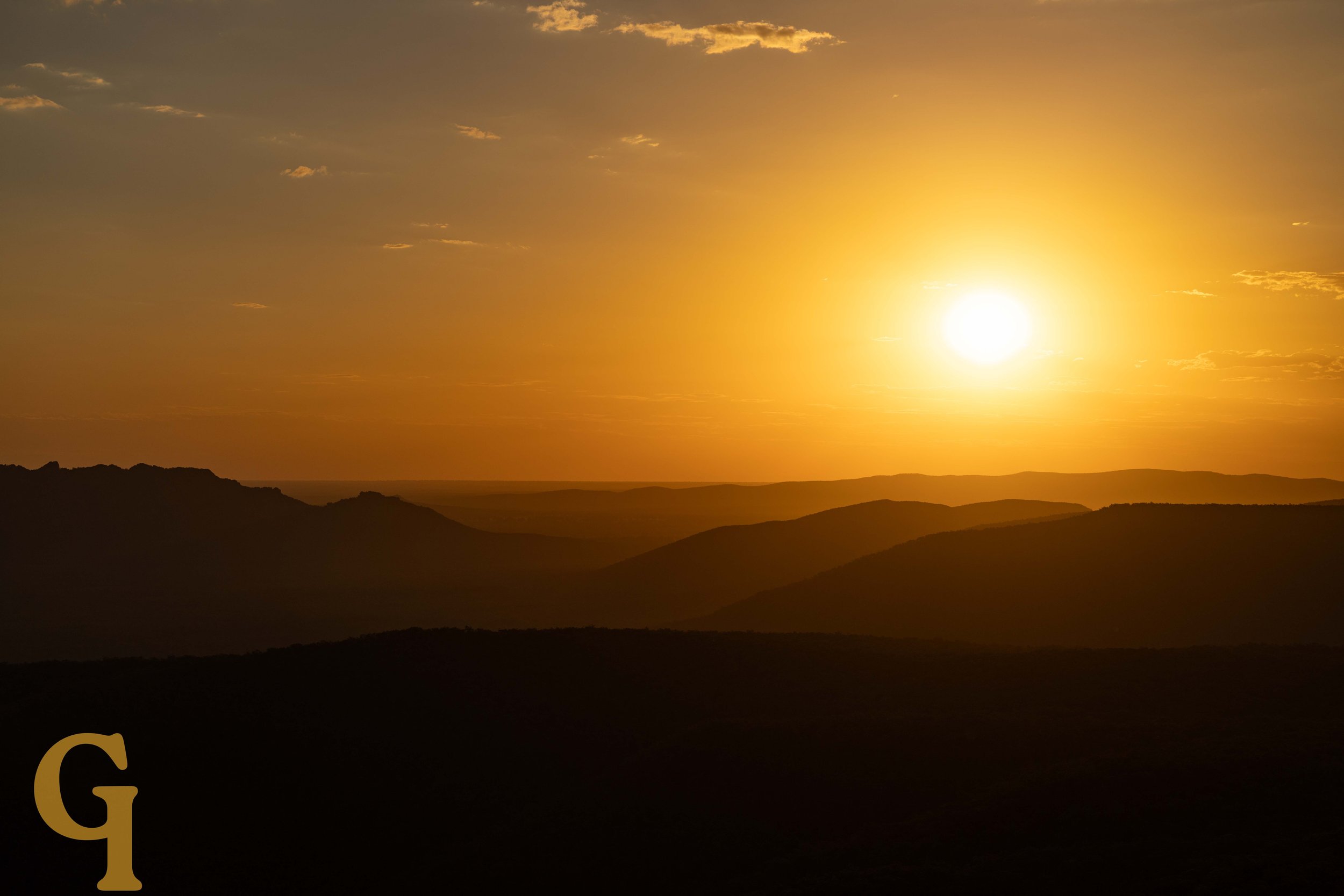 Sunset over mountains, with a golden sky and a few clouds.